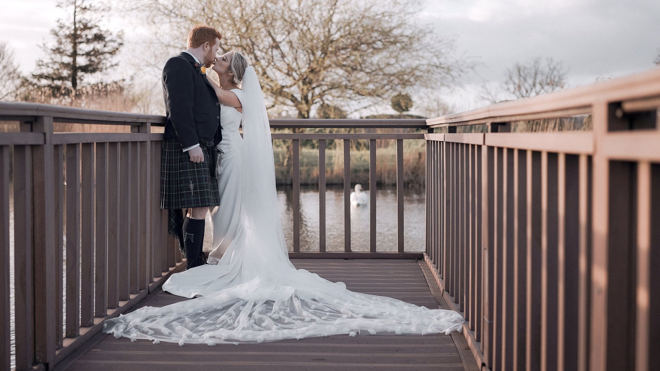 Bride and groom kissing on the wooden jetty by the lake with a swan at Ballymagarvey Village, Co. Meath