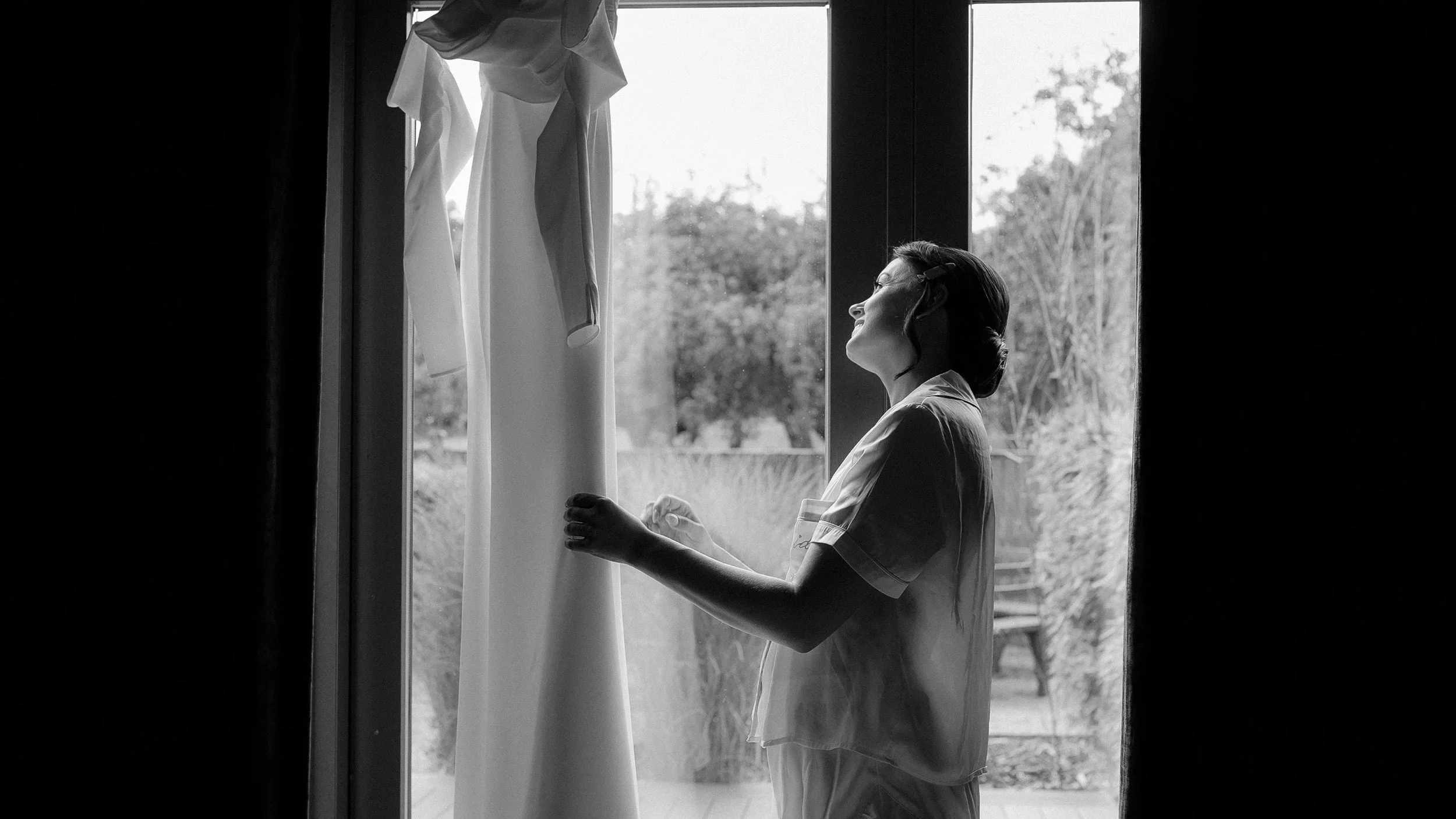 Black and white photo of the bride looking at her wedding dress hanging in the window at Farnham Estate