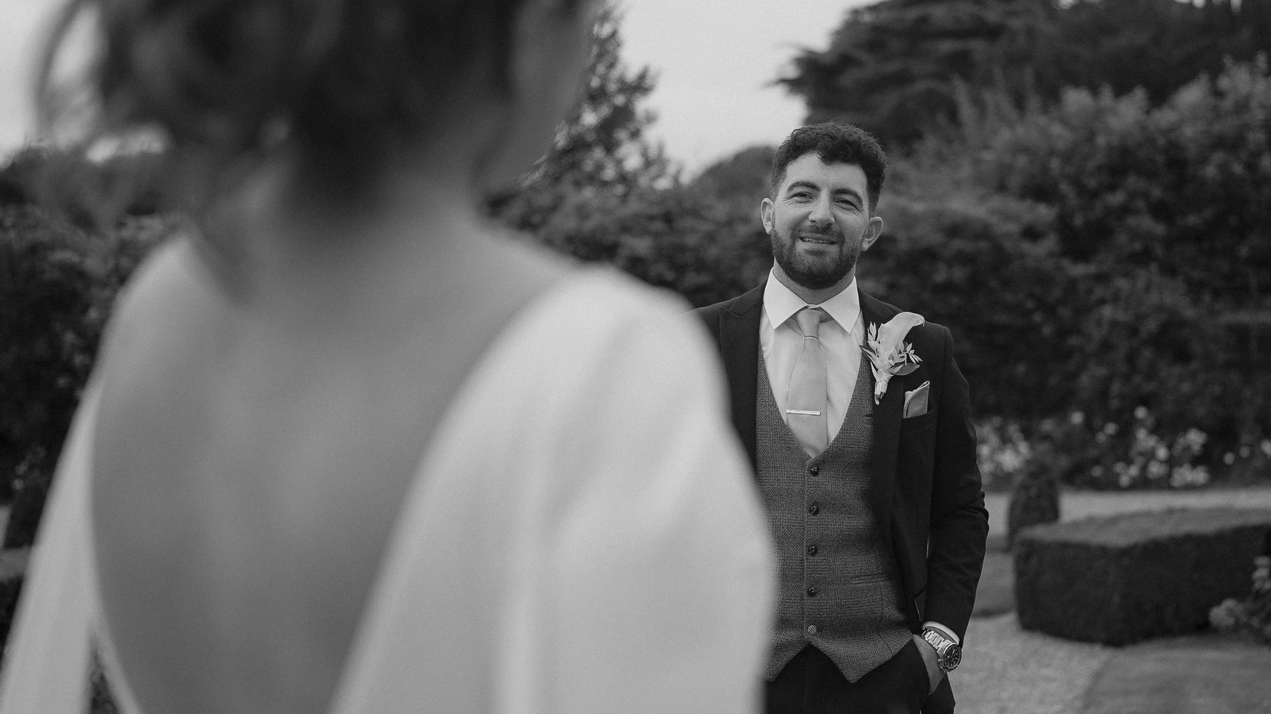 Groom in a tweed waistcoat smiling at his bride in the gardens of Kilkea Castle, black and white