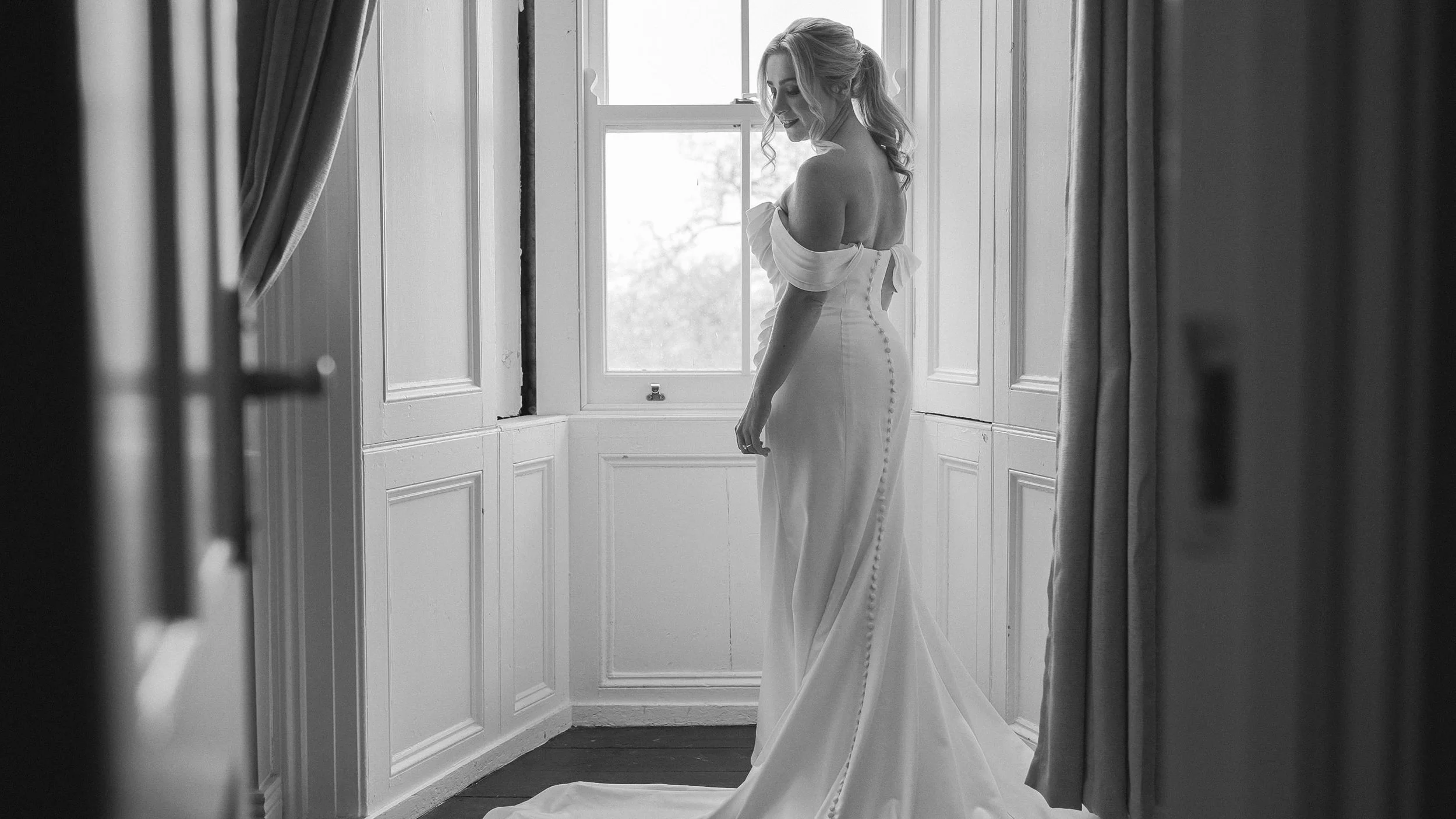 Black and white photo of the bride in her off-the-shoulder gown by a sash window at Ballymagarvey Village, Co. Meath