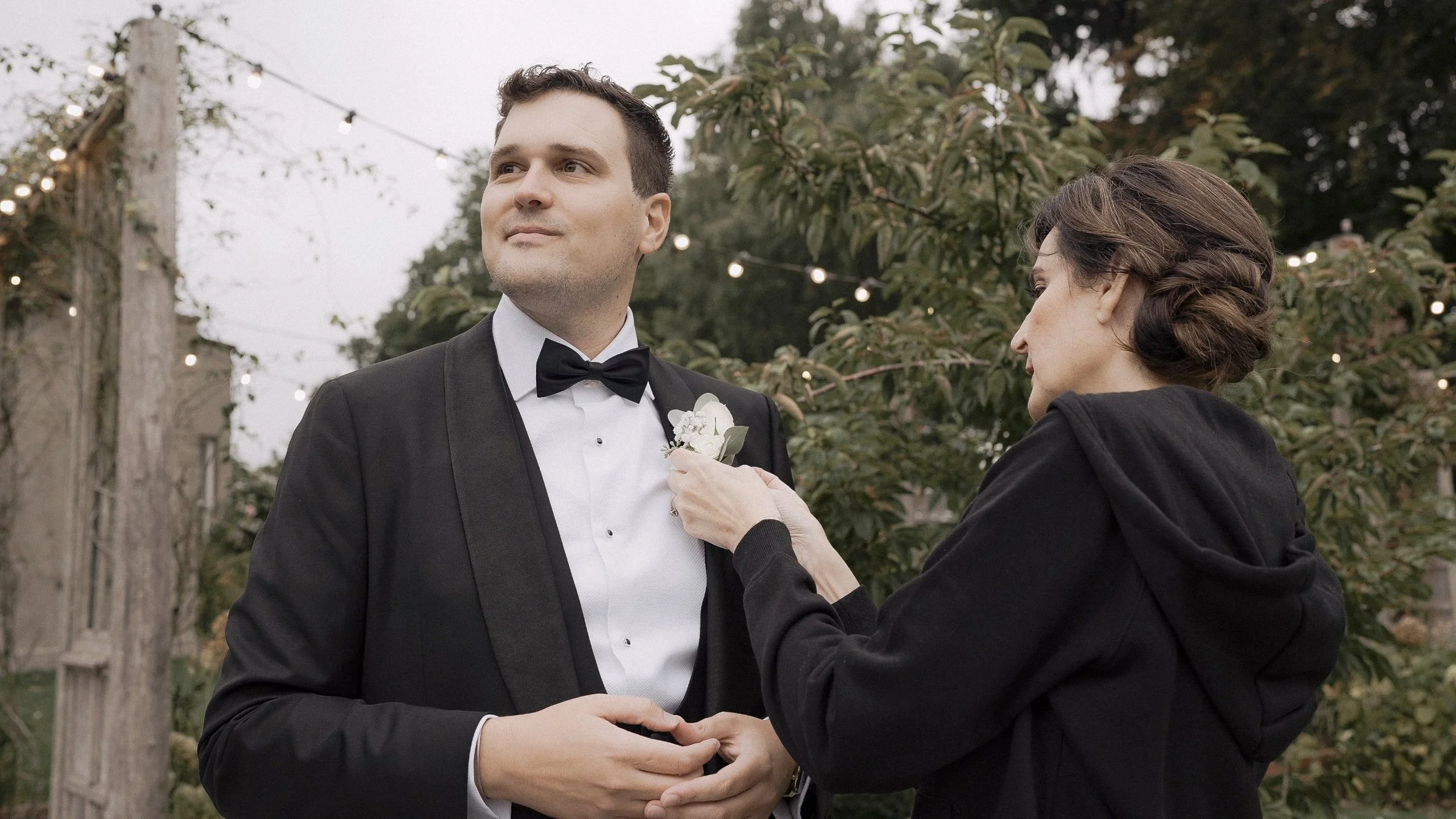 Groom having his buttonhole pinned on under festoon lights in the garden at Tinakilly House