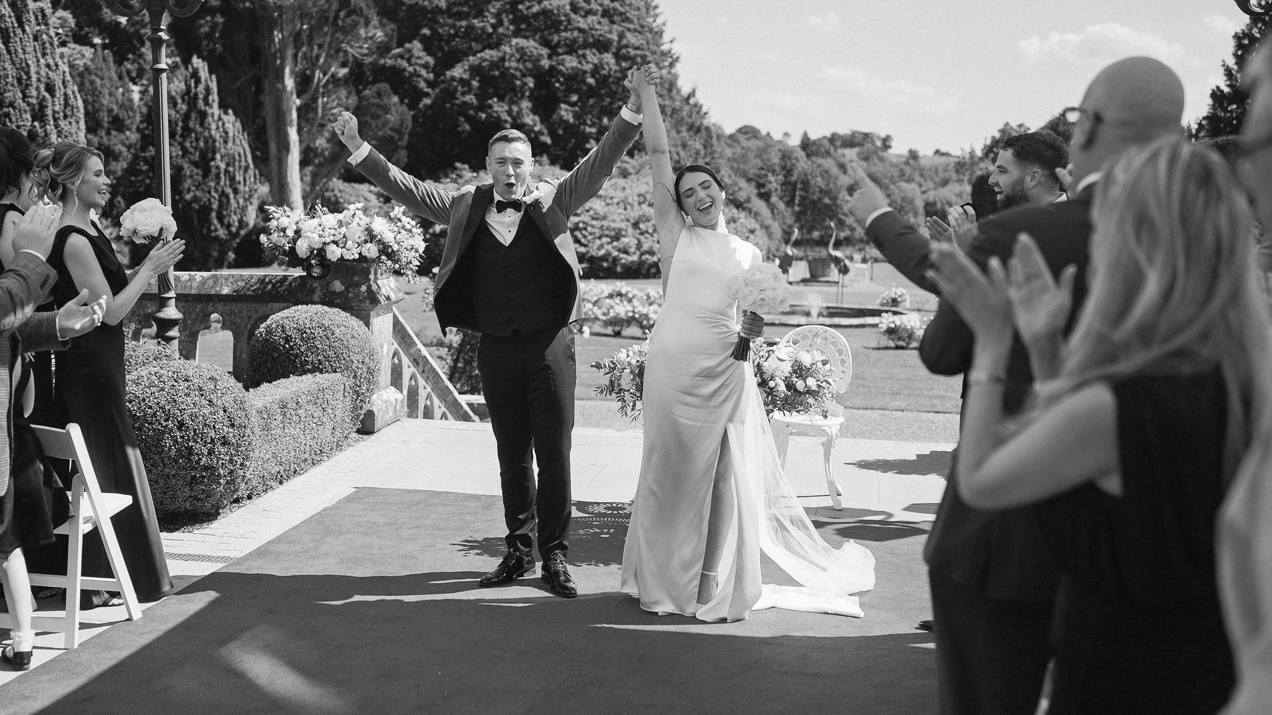 Newlyweds raising their arms in celebration as guests cheer after the outdoor ceremony at Markree Castle, black and white