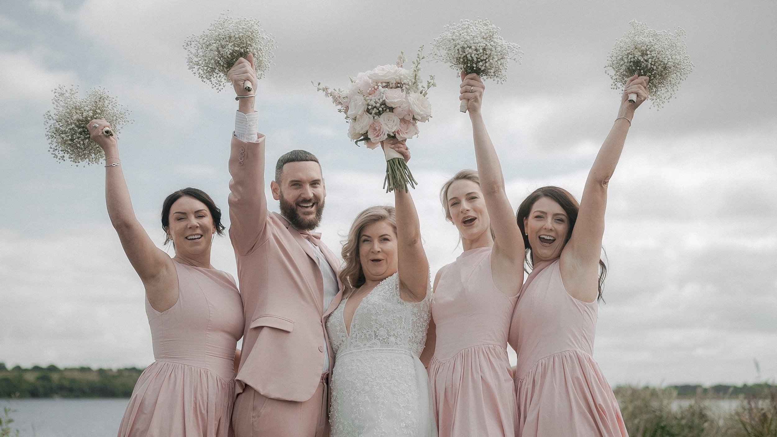 Bride and bridal party in blush pink raising their bouquets by Lough Ree at Glasson Lakehouse