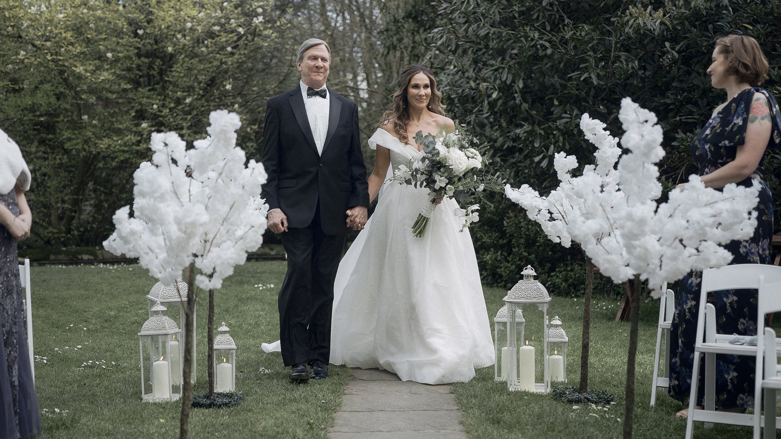 Bride walking down the garden aisle with her father between white blossom trees and lanterns at Waterford Castle