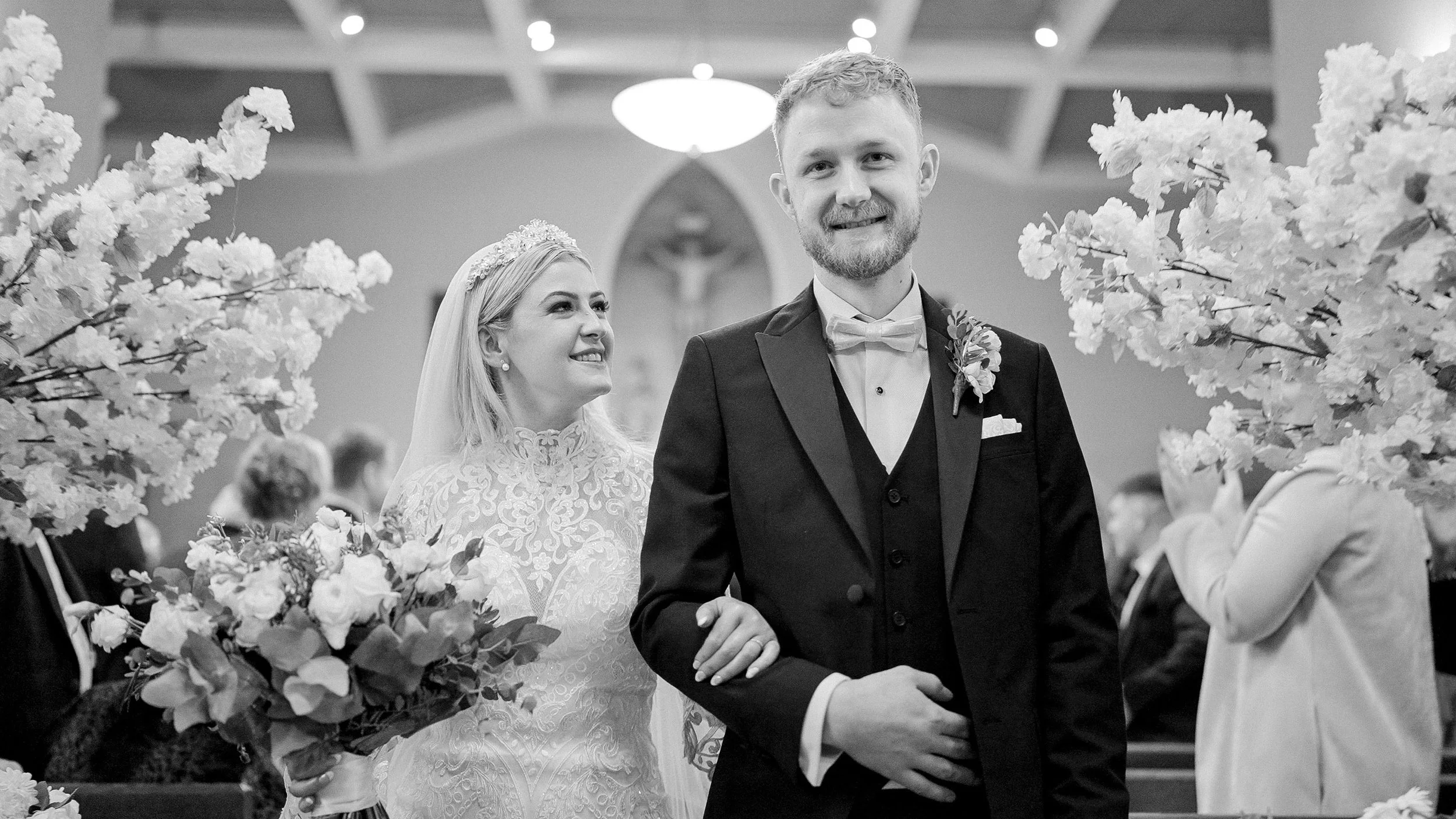 Black and white photo of Hannah and Francis walking down the aisle lined with blossom trees at St Joseph's Church, Milltown Malbay