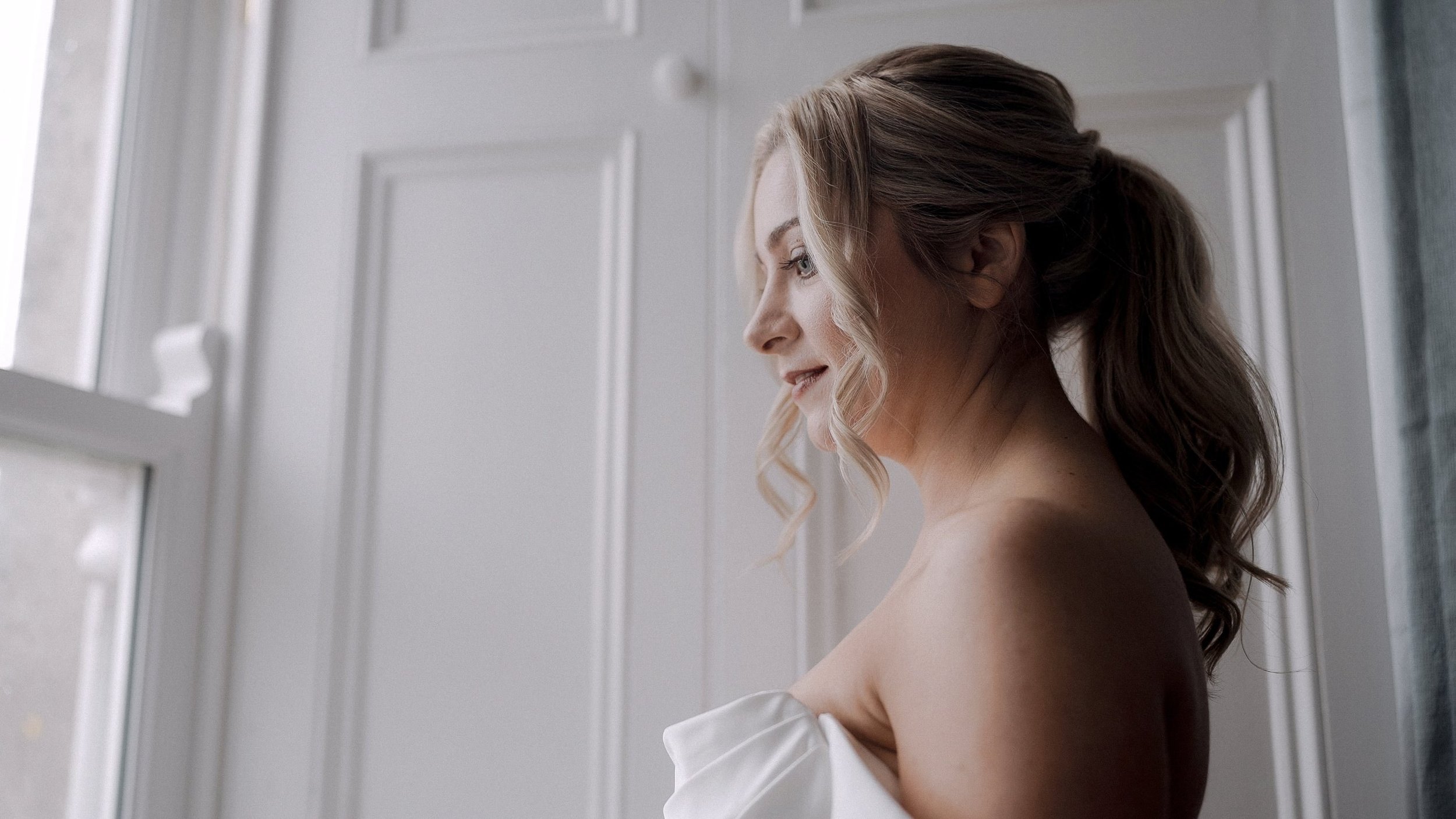Bride with a curled ponytail smiling by the window at Ballymagarvey Village, Co. Meath