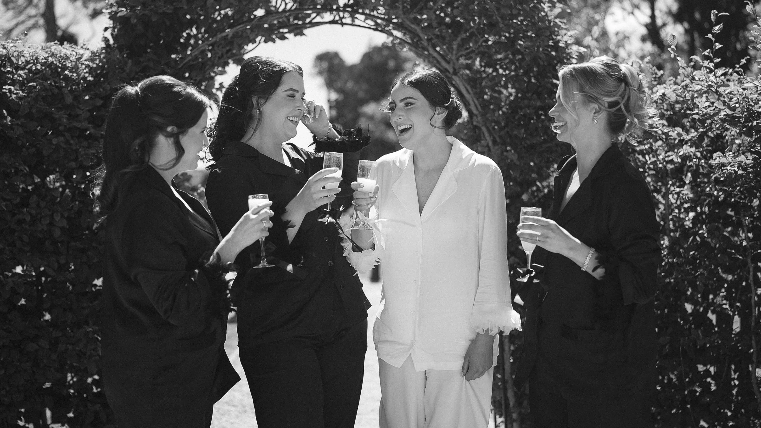 Bride in white pyjamas laughing with bridesmaids in black pyjamas and champagne in the garden at Markree Castle, black and white