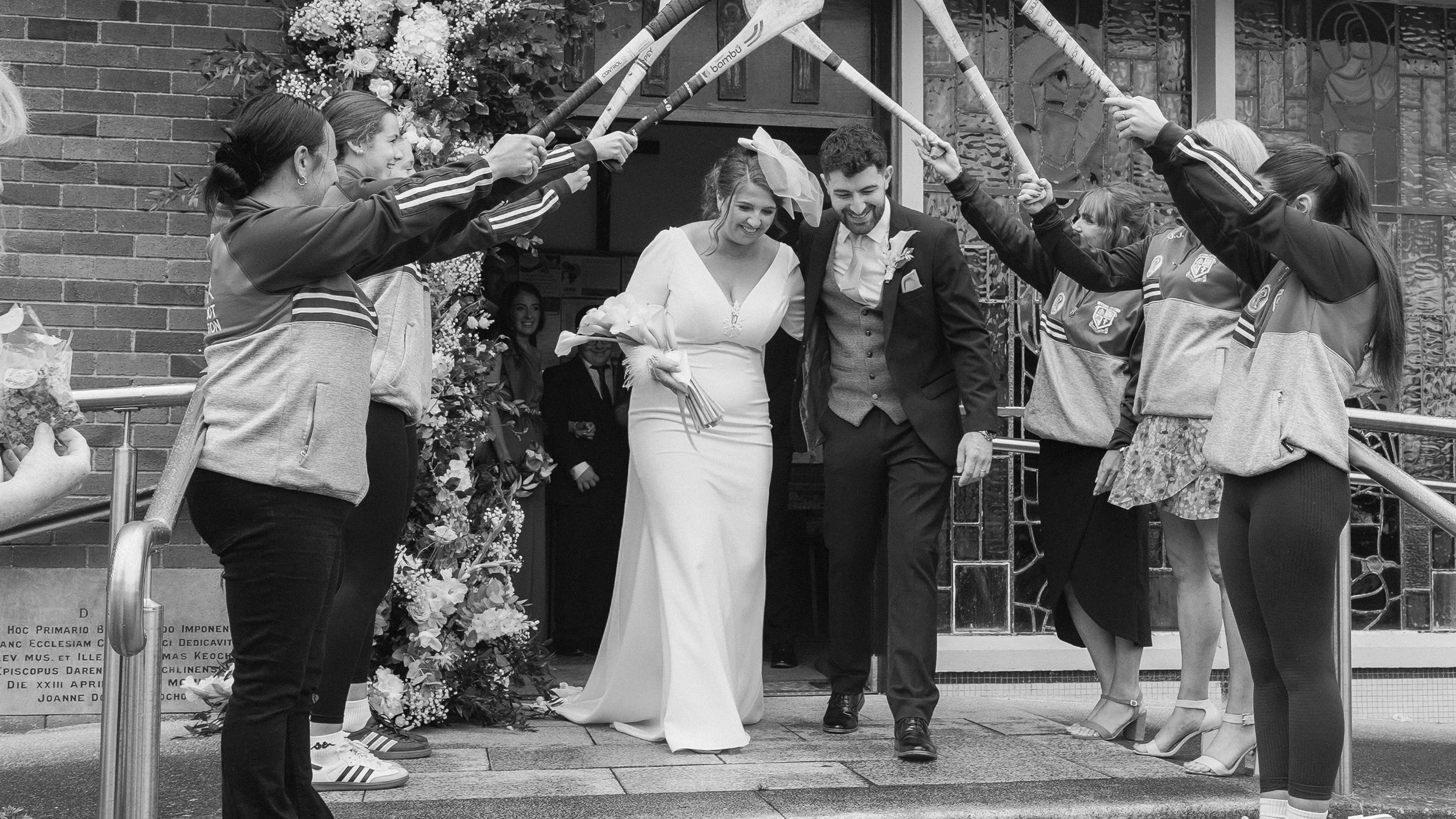 Bride and groom walking under a guard of honour of raised hurleys outside Christ the King Church, Cooleragh, black and white