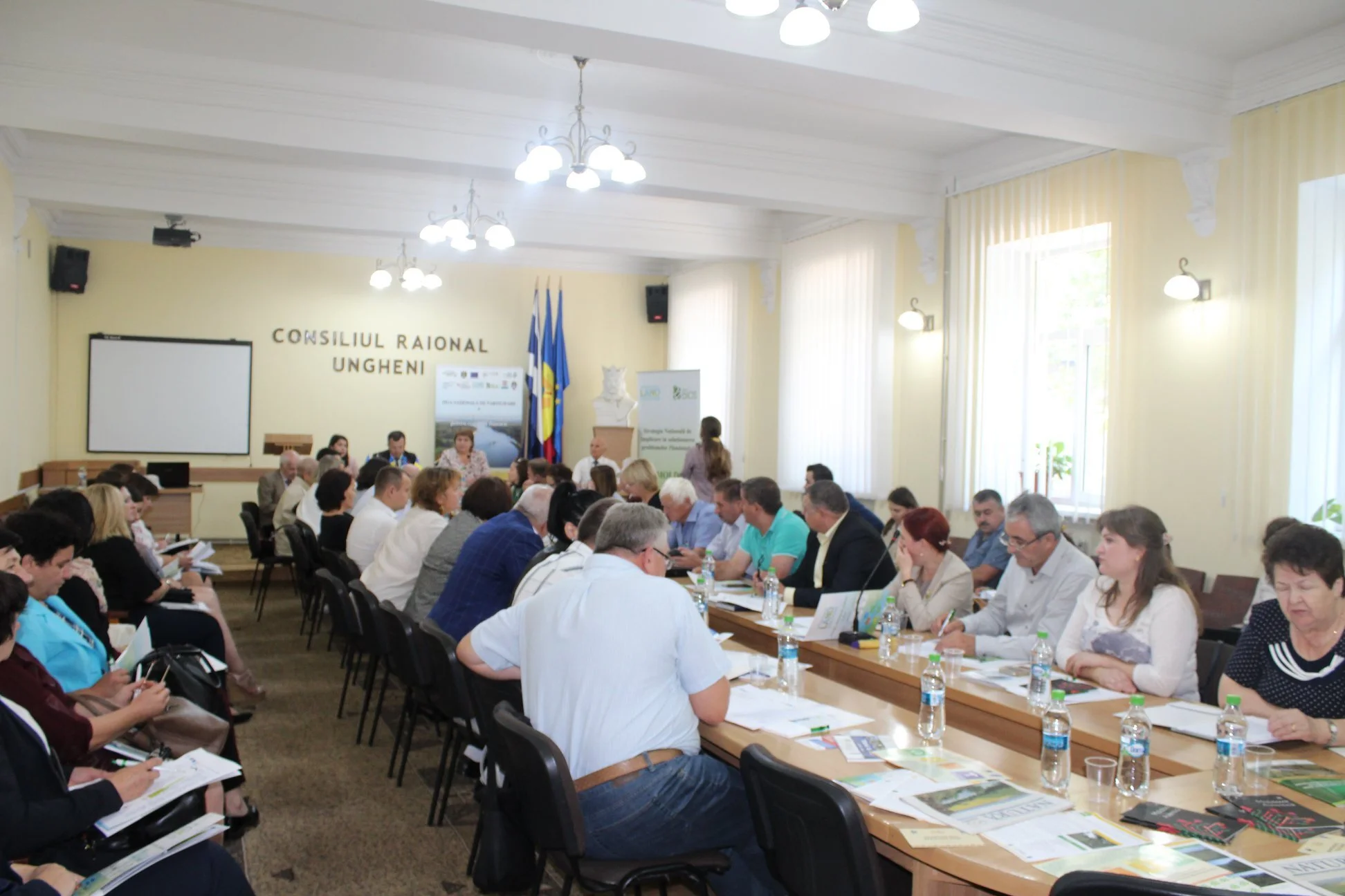People attending a meeting or conference in a large, well-lit room with cream walls and large windows. Participants are seated around a long U-shaped table with documents and water bottles. A few flags and a sign reading 'CONSILIUL RATIONA UNGHENI' are visible at the front.