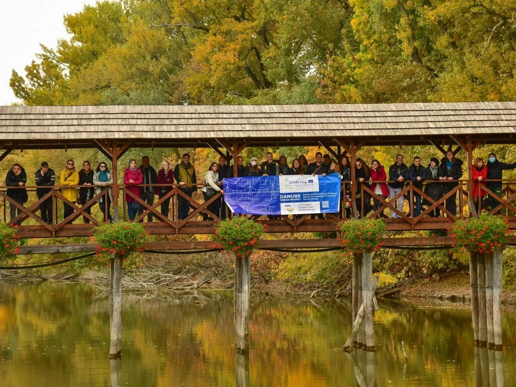 A group of people standing on a wooden bridge over a river, with trees in fall colors in the background, holding banners and wearing masks.