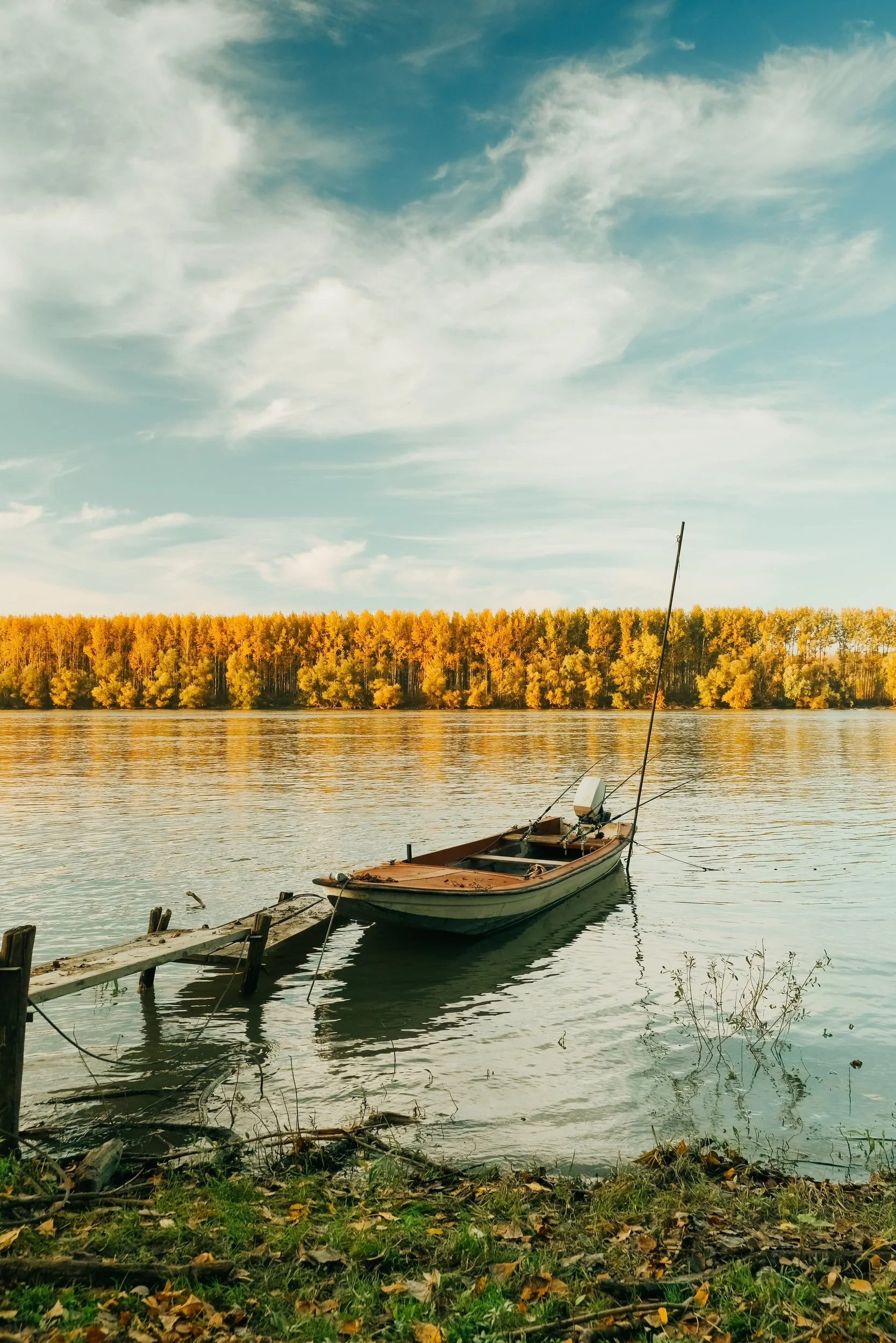 A boat tied to a wooden dock on a calm lake with an autumn-colored tree line and a partly cloudy sky in the background.