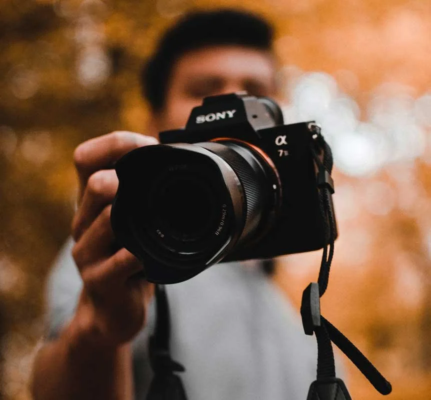 Person holding a Sony camera, pointing it toward the viewer, blurred background with warm lighting.
