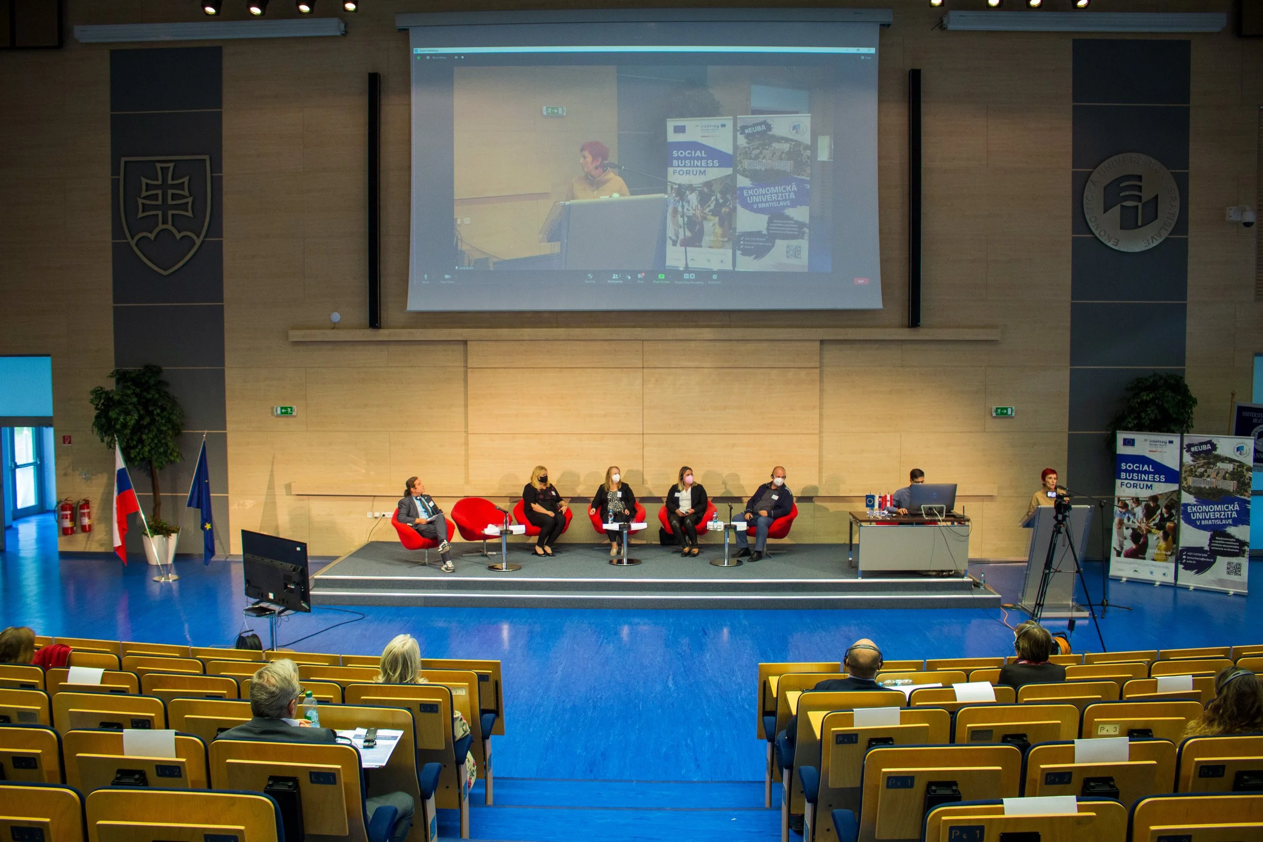 A panel of six people wearing face masks sitting on stage in front of a large screen displaying a video call. The event is a Social Business Forum at a university auditorium with an audience seated in front.