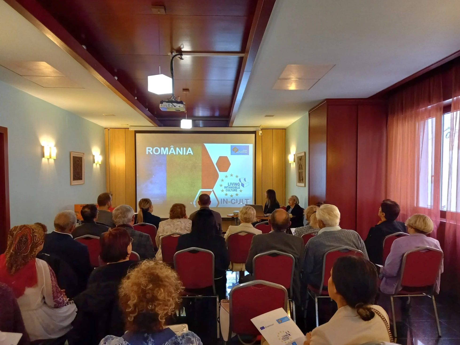 A conference room with people attending a presentation on Romania, with a screen displaying Romanian flag and European Union symbols.