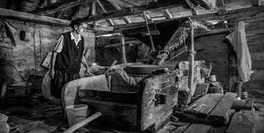 A woman in traditional clothing standing in an old workshop or barn with an antique woodworking or crafting machine.