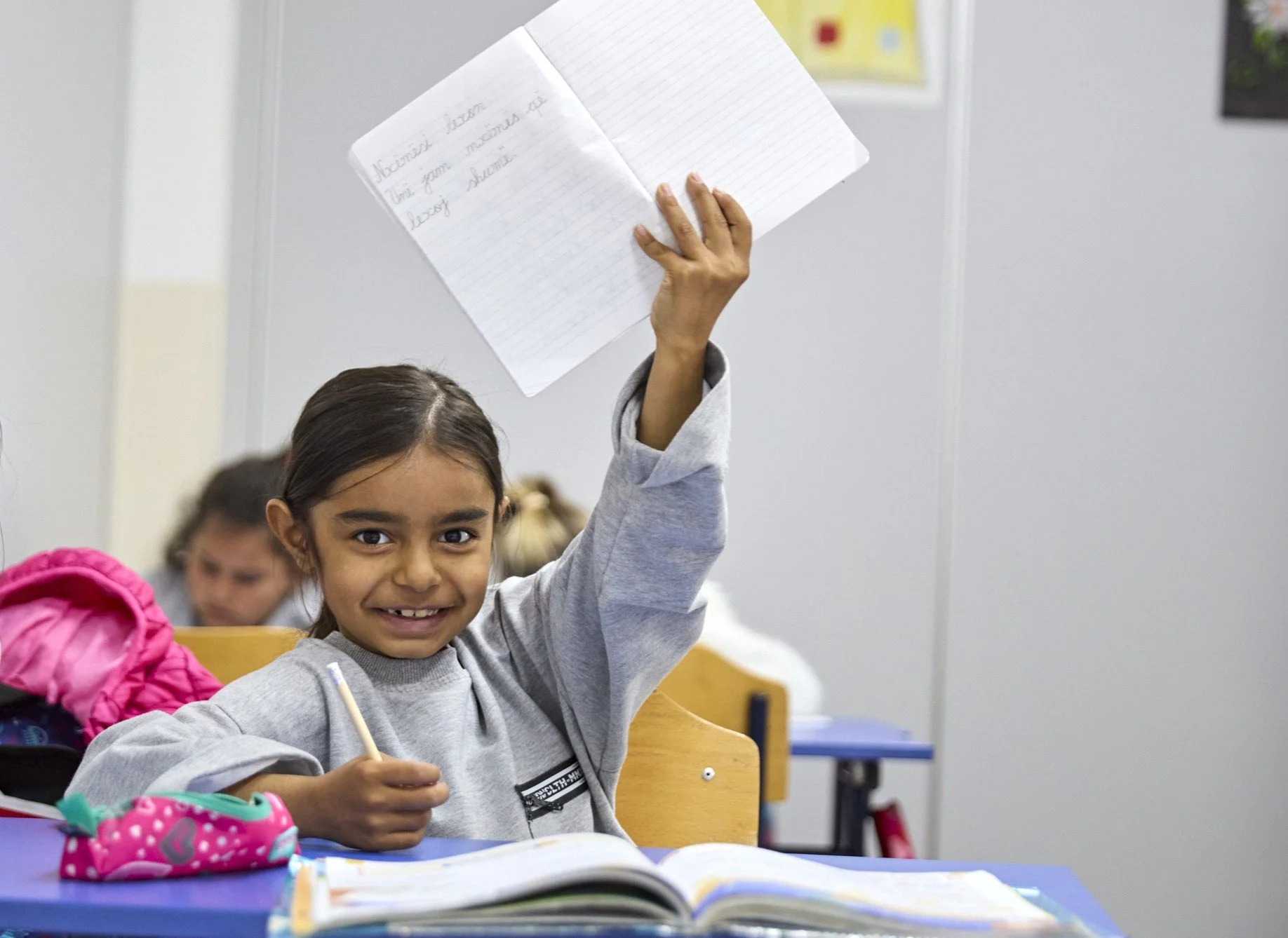 A young girl in a classroom raising her hand, holding a notebook, with open books on her desk and other students in the background.