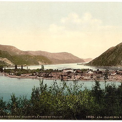 A watercolor painting of a lakeside village set between mountains with greenery in the foreground and a body of water reflecting the sky.