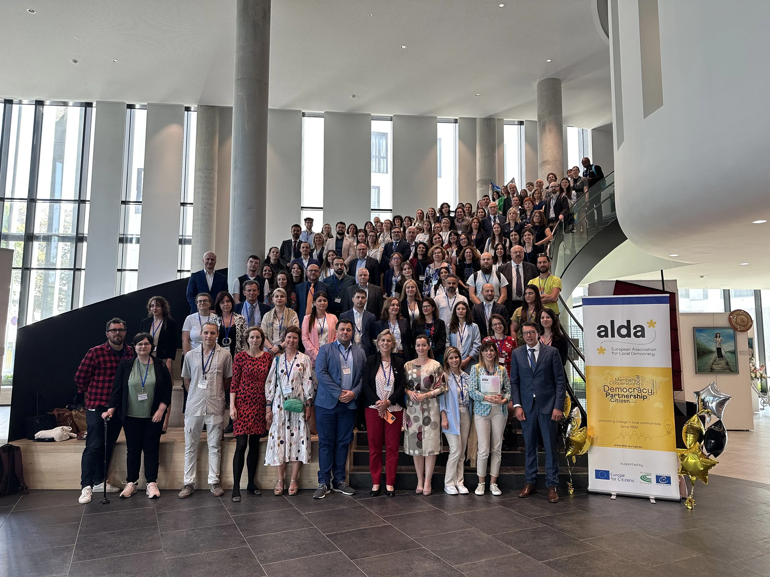 Group of diverse people gathered on a staircase at a conference or event, with a banner for ALDA, European Association for Local Democracy, in a modern building with large windows.
