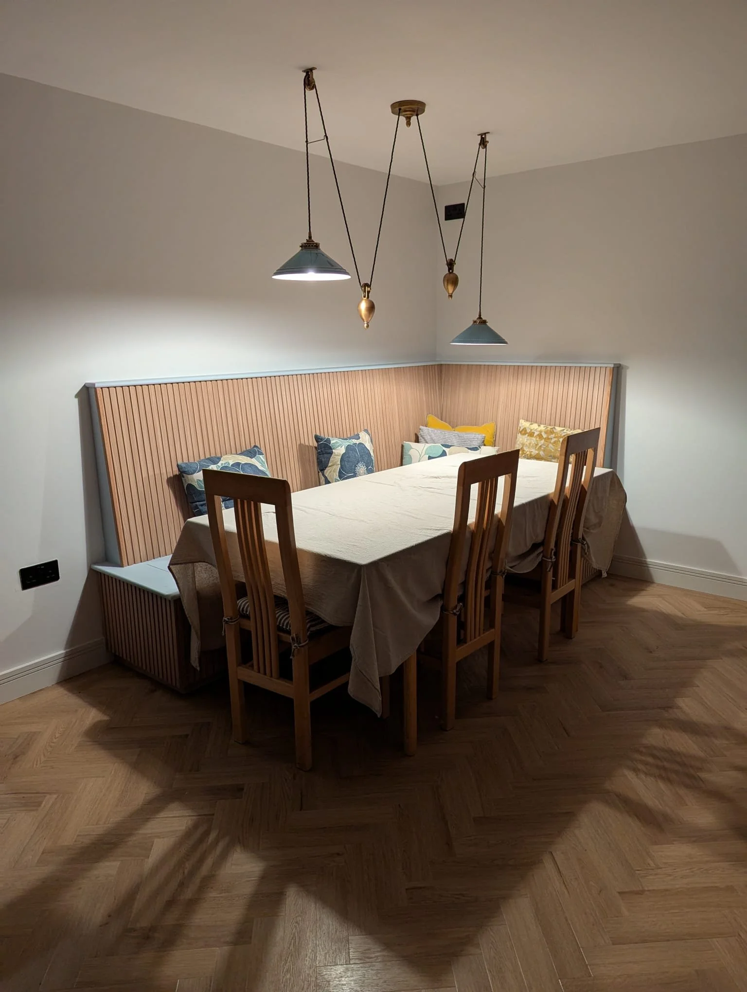 Dining room with wooden table, chairs, and a padded headboard back wall. Three hanging pendant lights above the table. Cushions on the seating area with colorful patterns. Hardwood floor.