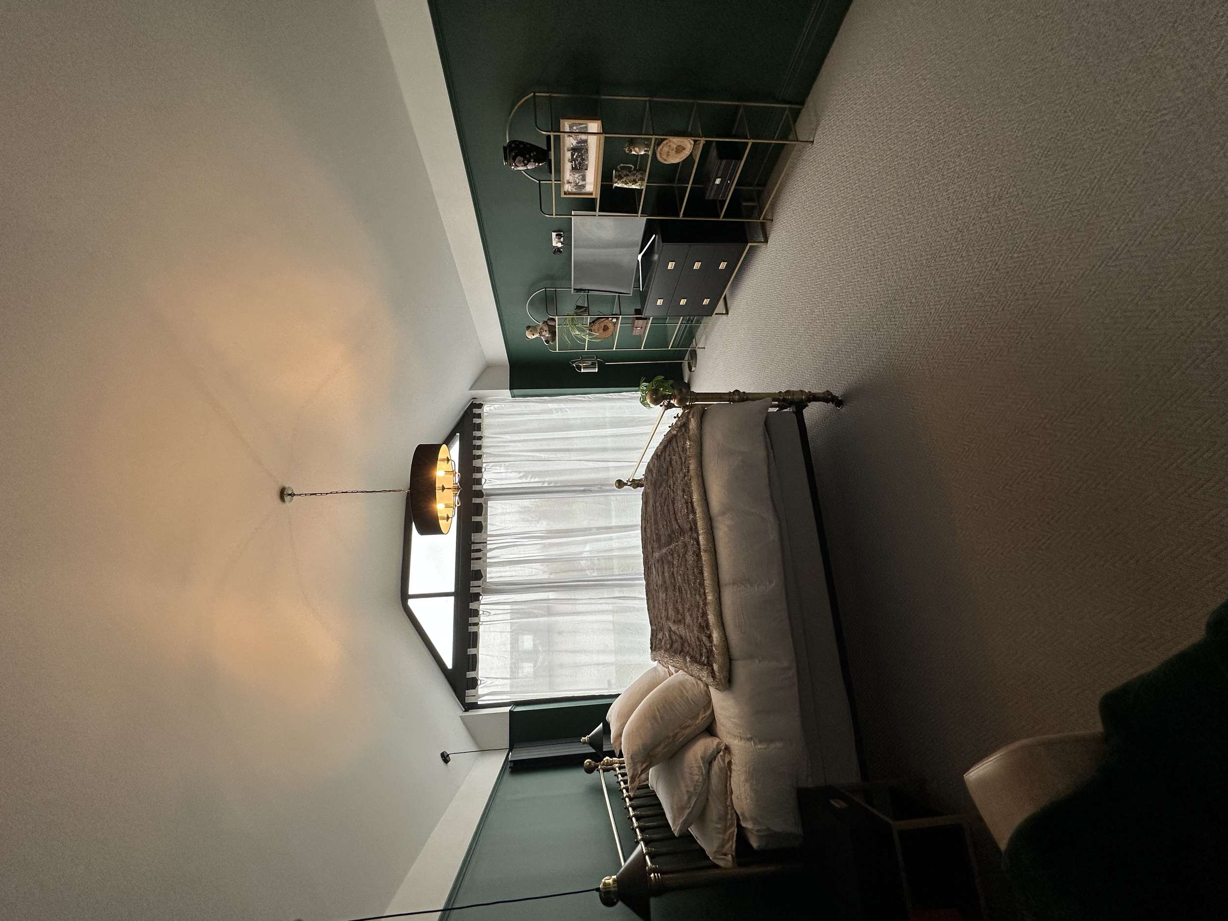 A bedroom with a vintage bed, plush pillows, and a brown blanket. A black and gold shelving unit with decorative items rests against a green accent wall. A large window with sheer white curtains allows natural light. A modern black and gold pendant l