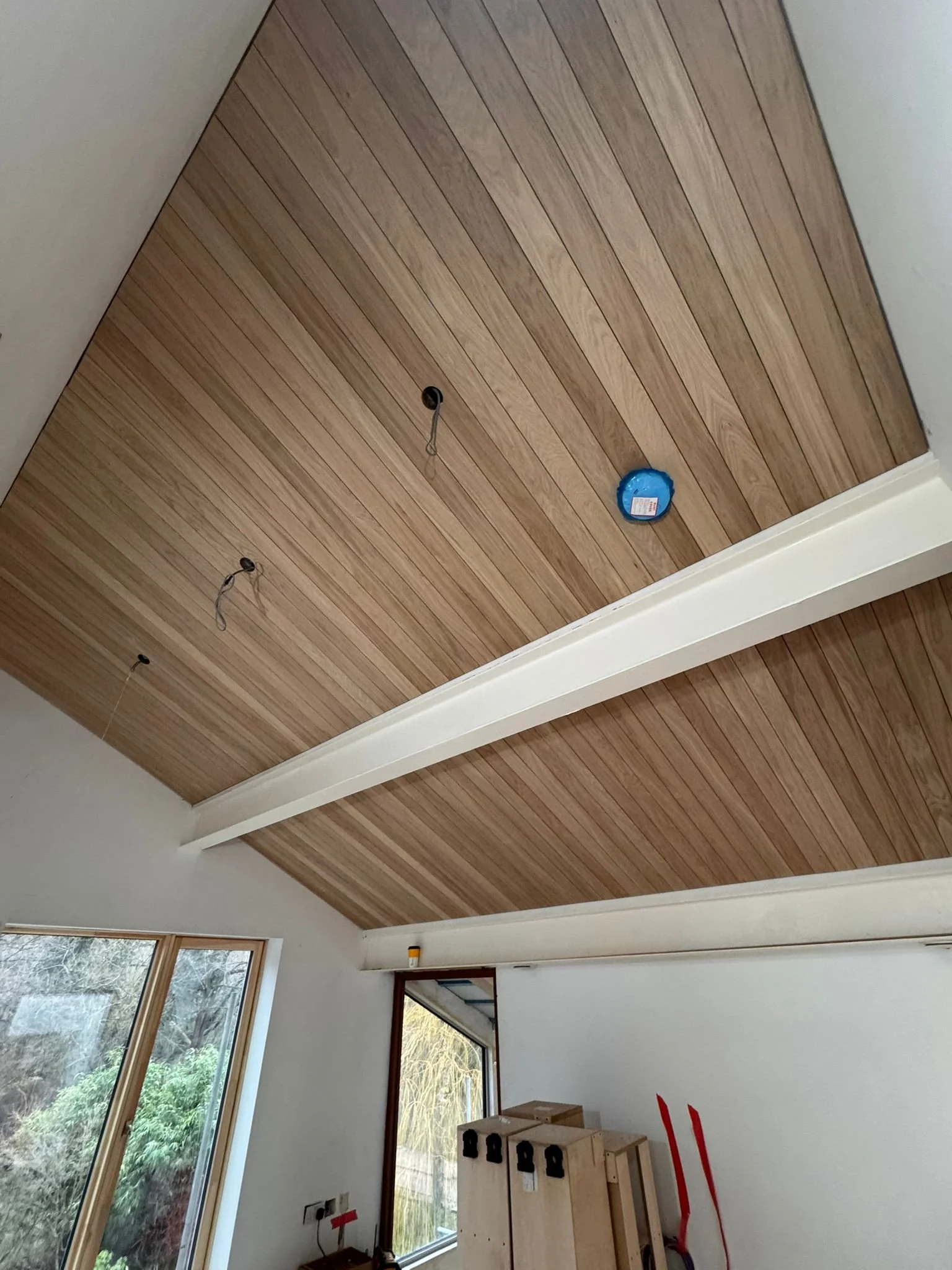 Interior room with a wood-paneled ceiling, ceiling light fixtures not yet installed, and a window showing an outdoor view of trees and shrubs.