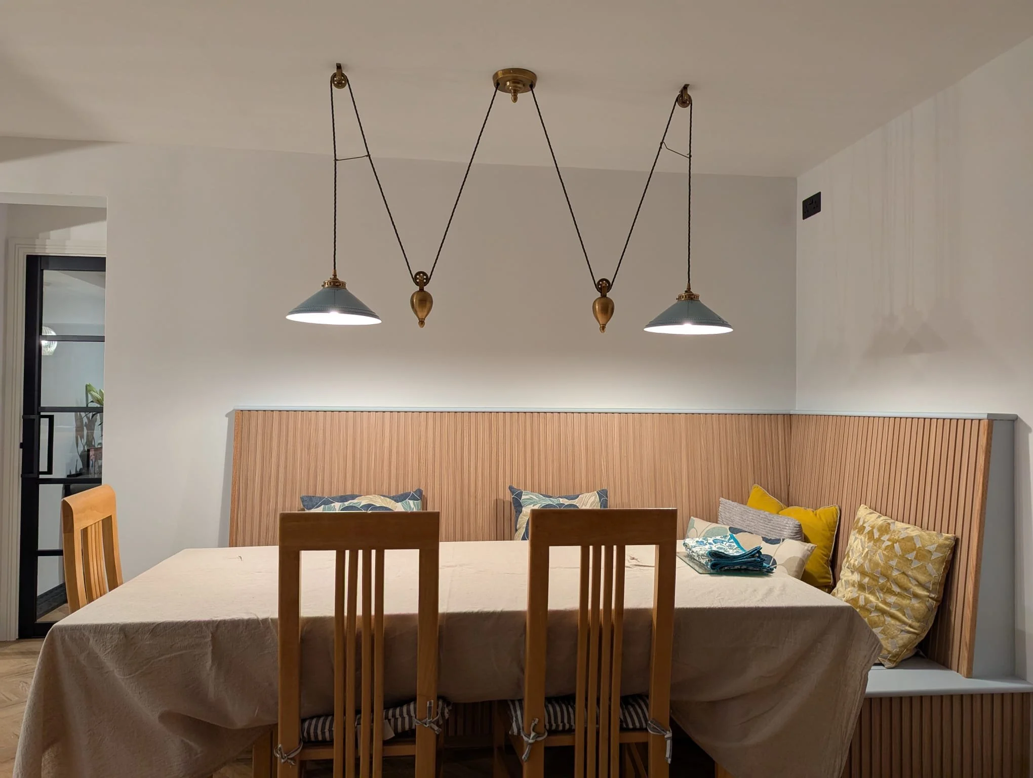 Dining room with a wooden table, beige tablecloth, six wooden chairs, a beige and yellow pillow arrangement on a built-in wooden bench, and a modern black hanging light fixture with three lights.