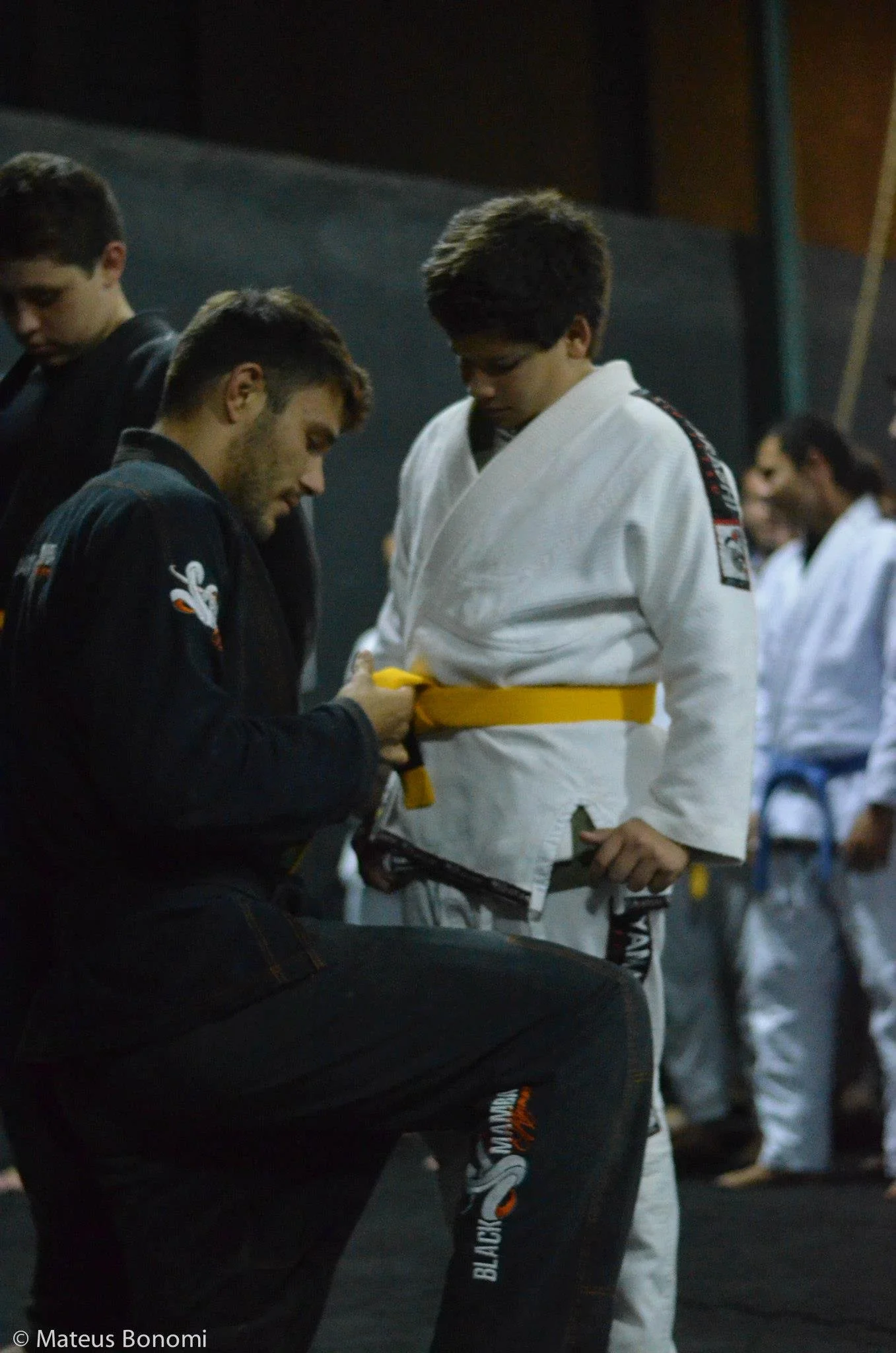 A young man receiving a yellow belt in Brazilian Jiu-Jitsu from an instructor during a class or competition, with other practitioners in the background.