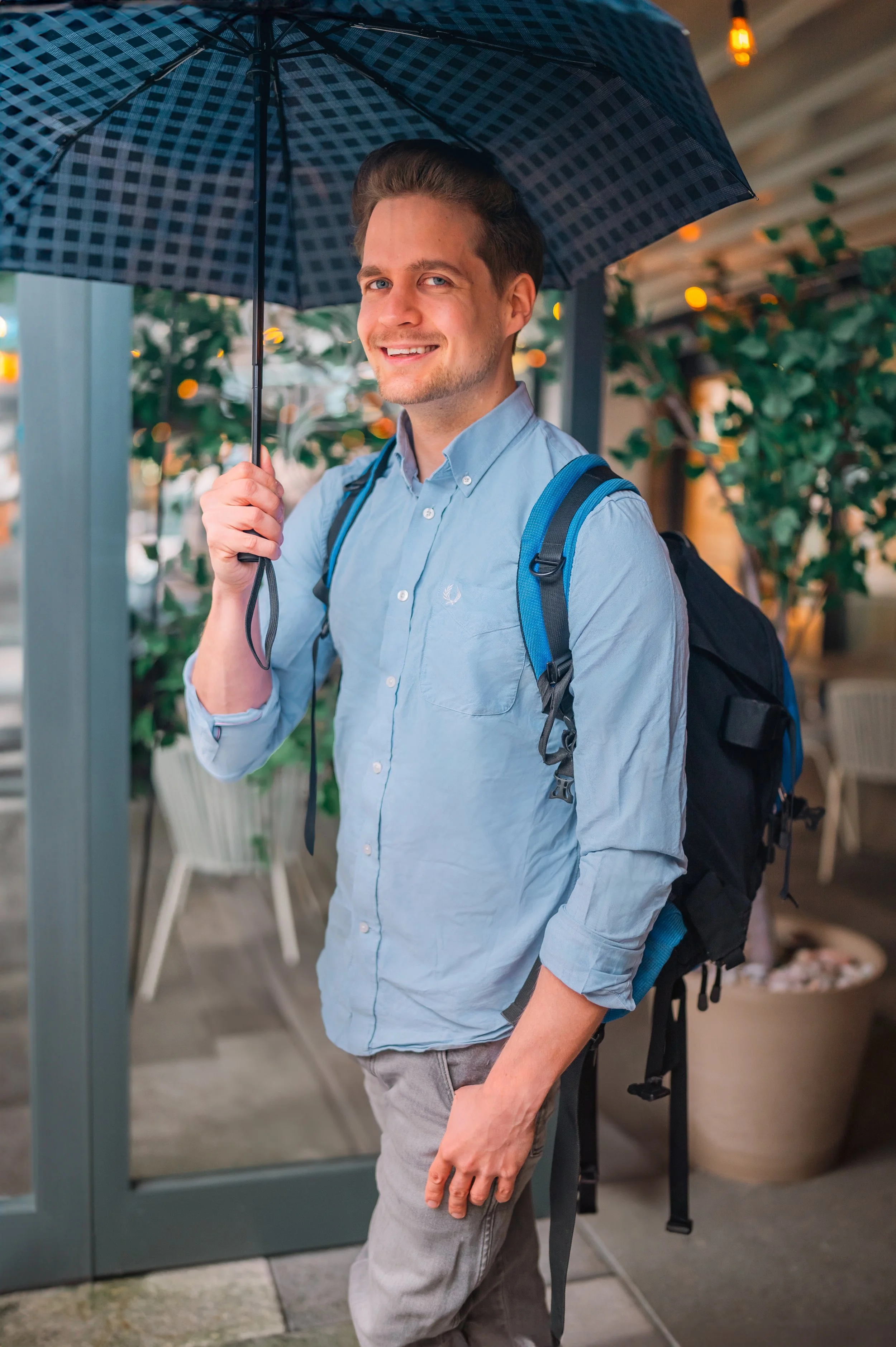 A smiling man with brown hair, wearing a light blue shirt and gray pants, holding an open chessboard with a rainbow umbrella over it, stands in front of an entrance with a glass door and plants.