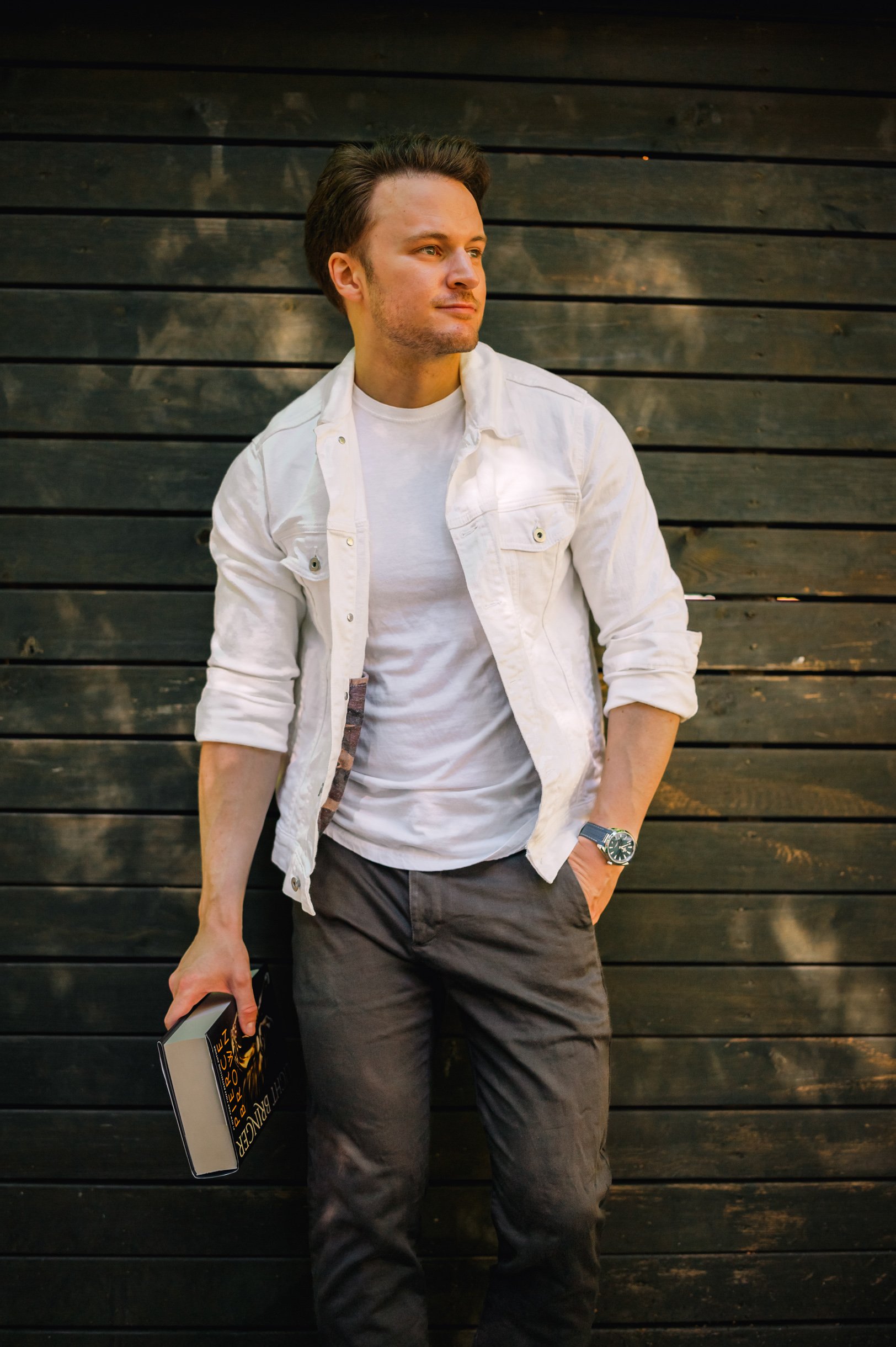 Young man standing against a dark wooden wall, wearing a white t-shirt and light denim jacket, with a book in his hand and looking to the side.