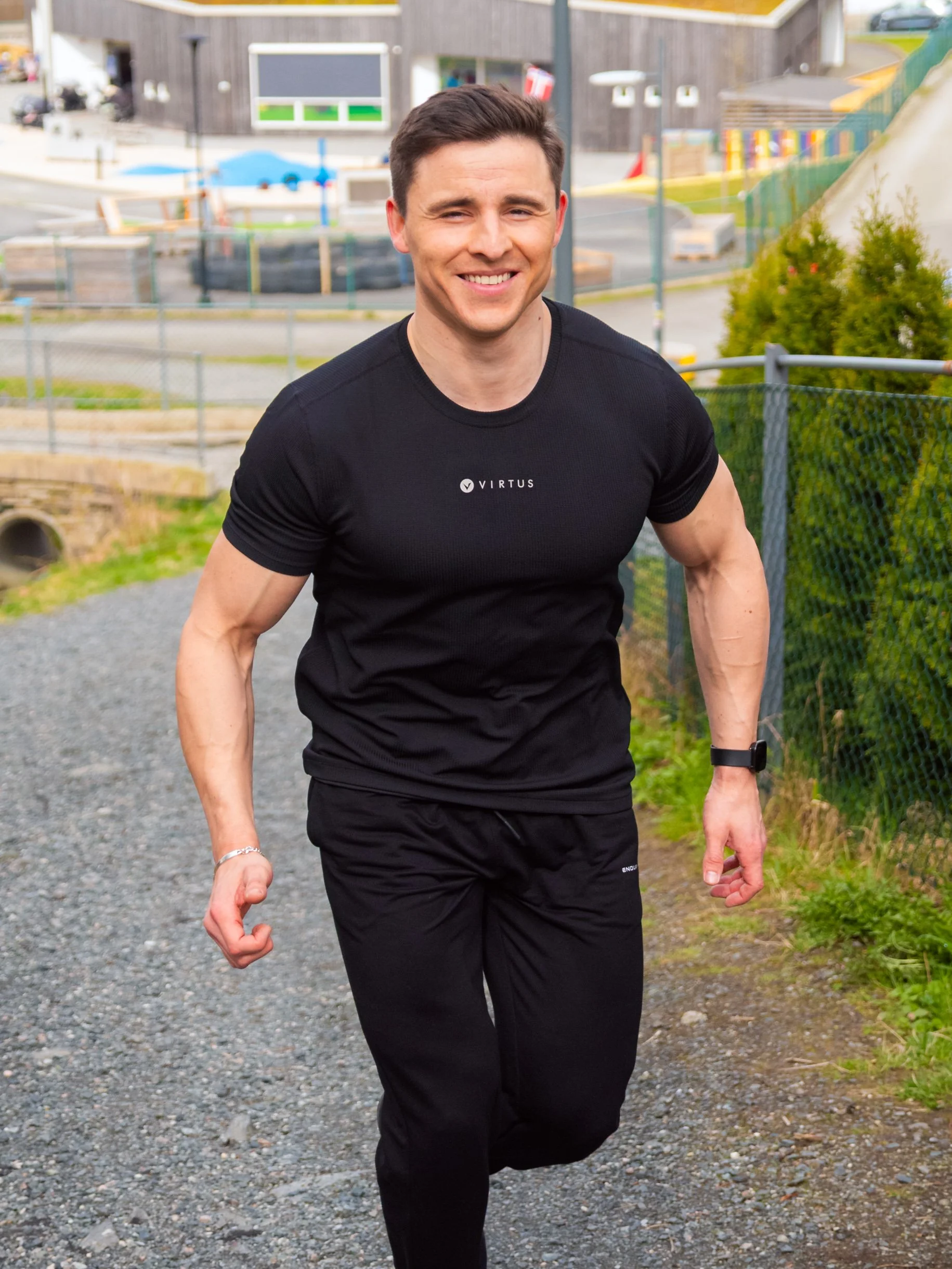 A man jogging outdoors on a dirt road, smiling. He has short brown hair and is wearing black tracksuits with the 'Virtus' logo.