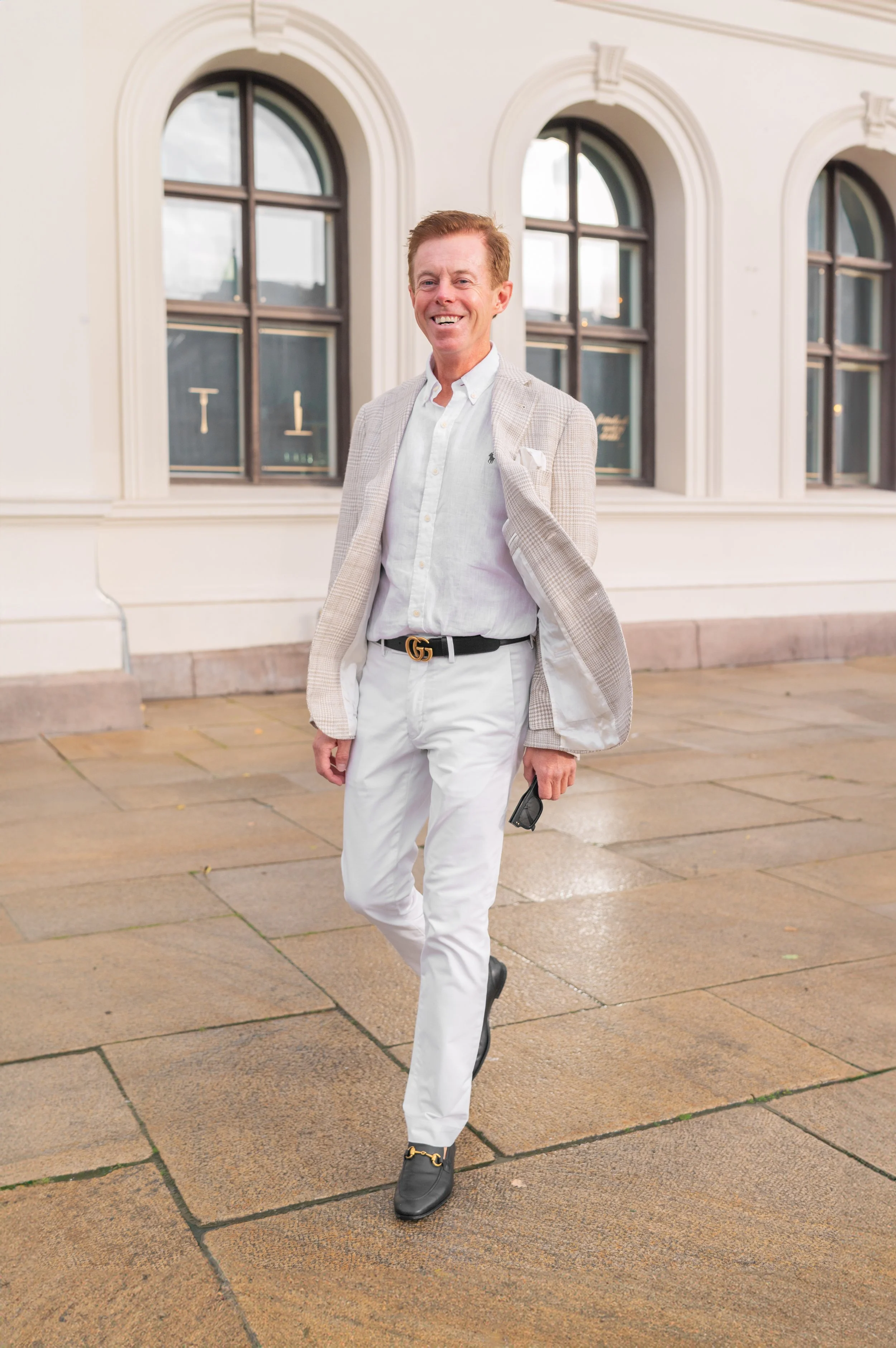 A man with red hair walking outdoors on a sidewalk, wearing a white shirt, white pants, black belt and black shoes, smiling. Background with a white building and large, arched windows.