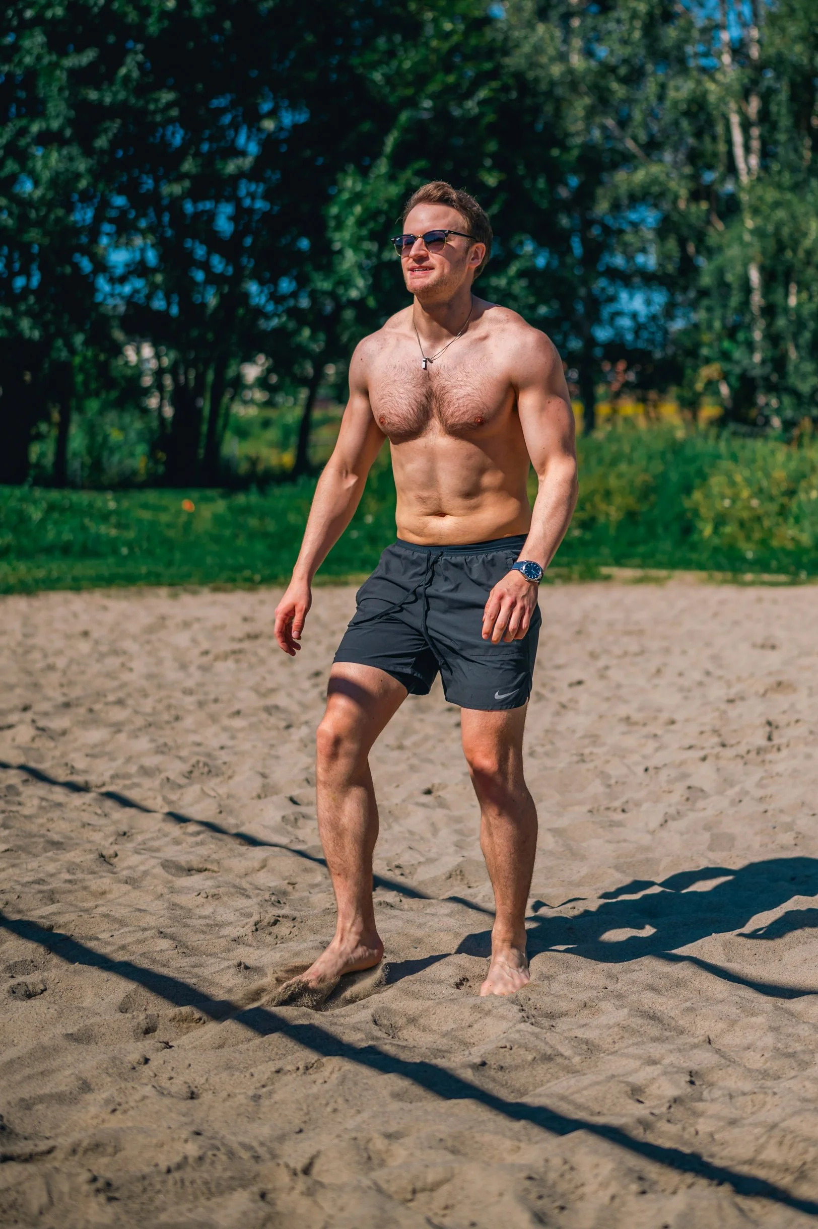 A shirtless man in black shorts walks on sand in a park with green trees.