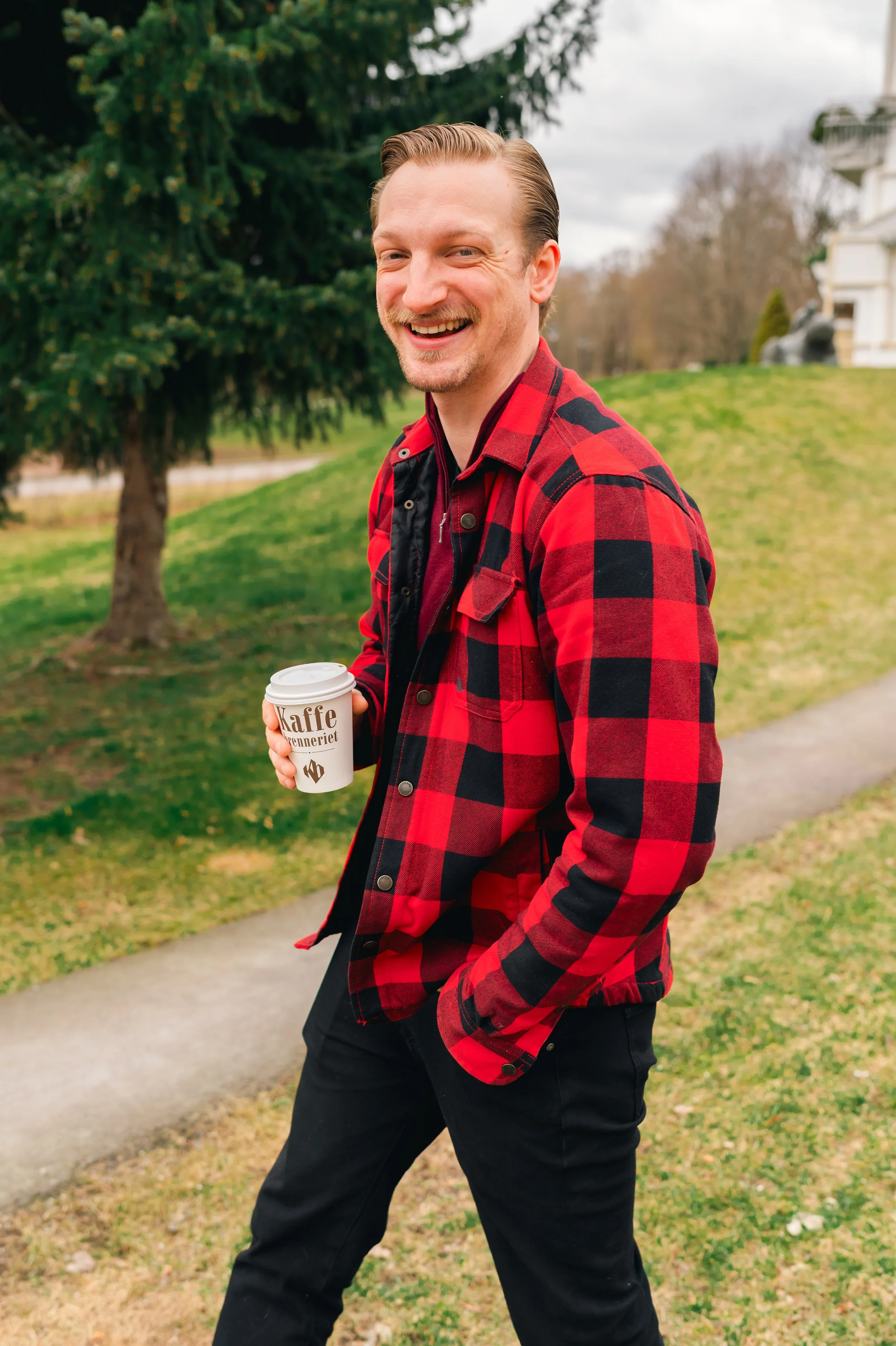 A man with fair skin, blond hair and a beard smiles while holding a cup of coffee, wearing a black and red checkered shirt and black pants, outdoors with green grass and trees in the background.