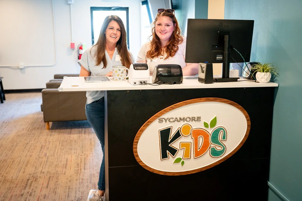 Two women standing behind a reception counter at Sycamore Kids, smiling at the camera in a brightly lit office space.