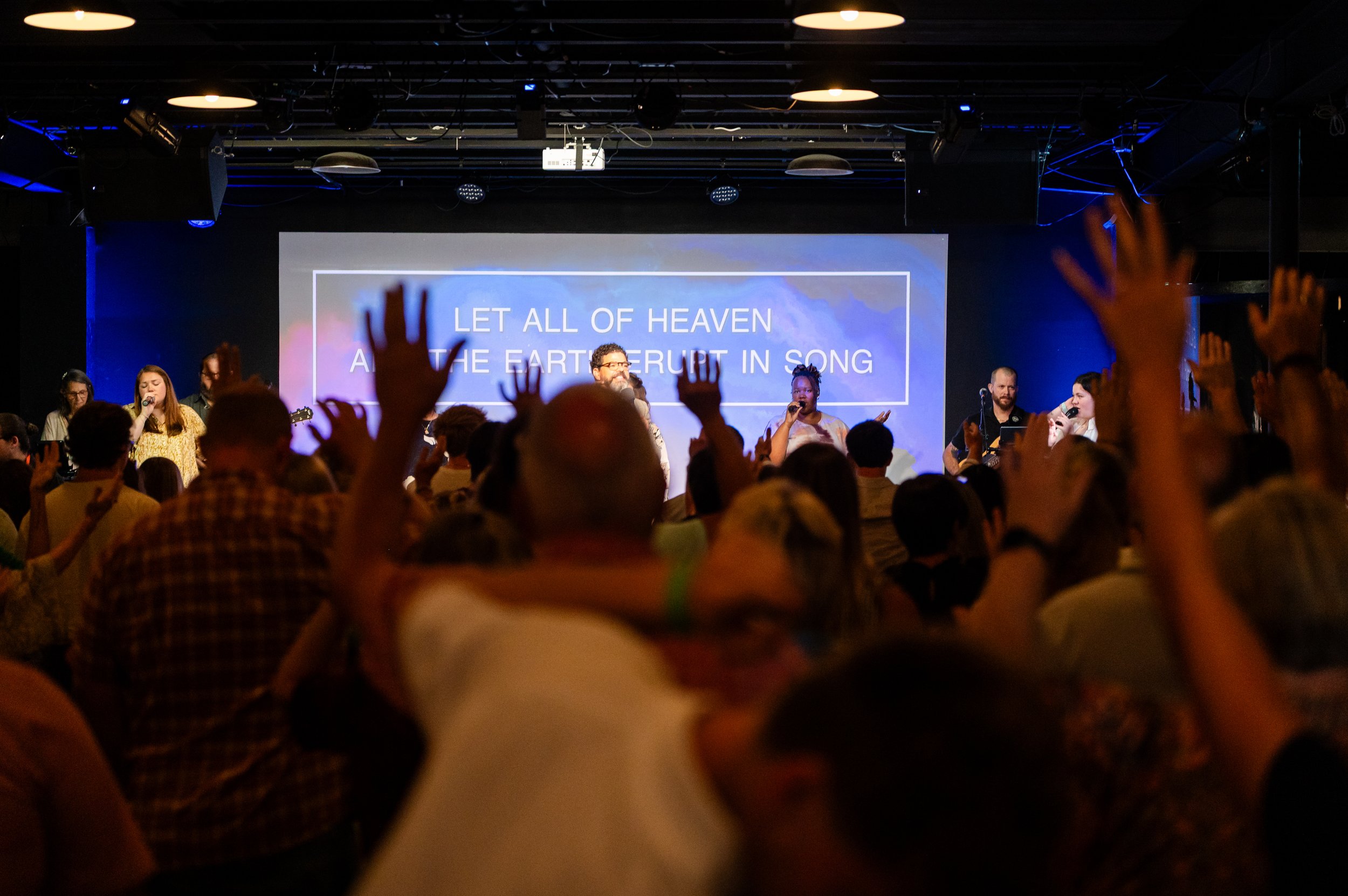 Crowd attending a church concert with a screen displaying song lyrics, and some raising their hands.