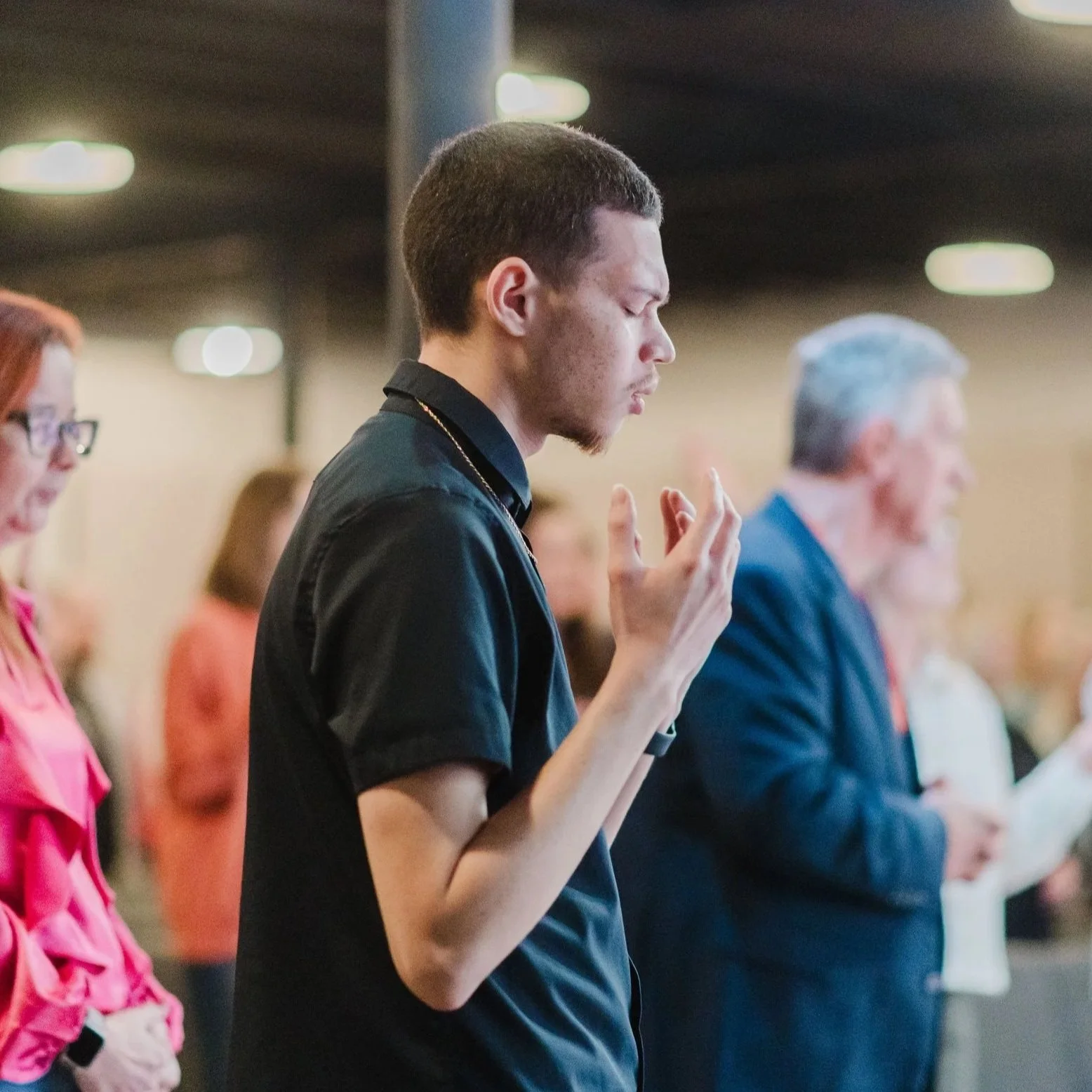 Two men hugging and smiling, one wearing a striped hoodie and baseball cap, the other in a denim shirt holding a paper, indoors.