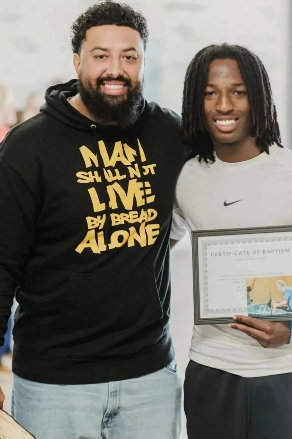 A man with a beard and a curly hairstyle standing next to a young man with dreadlocks, who is holding a certificate of baptism.