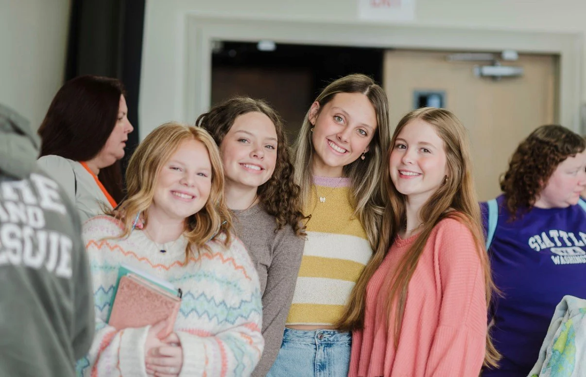 Group of five smiling teenage girls standing together indoors, posing for a photo.