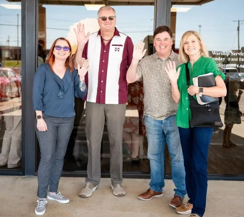 Four adults standing outside a glass door, waving at the camera, smiling. Two women and two men, casually dressed, with a parking lot visible in the background.
