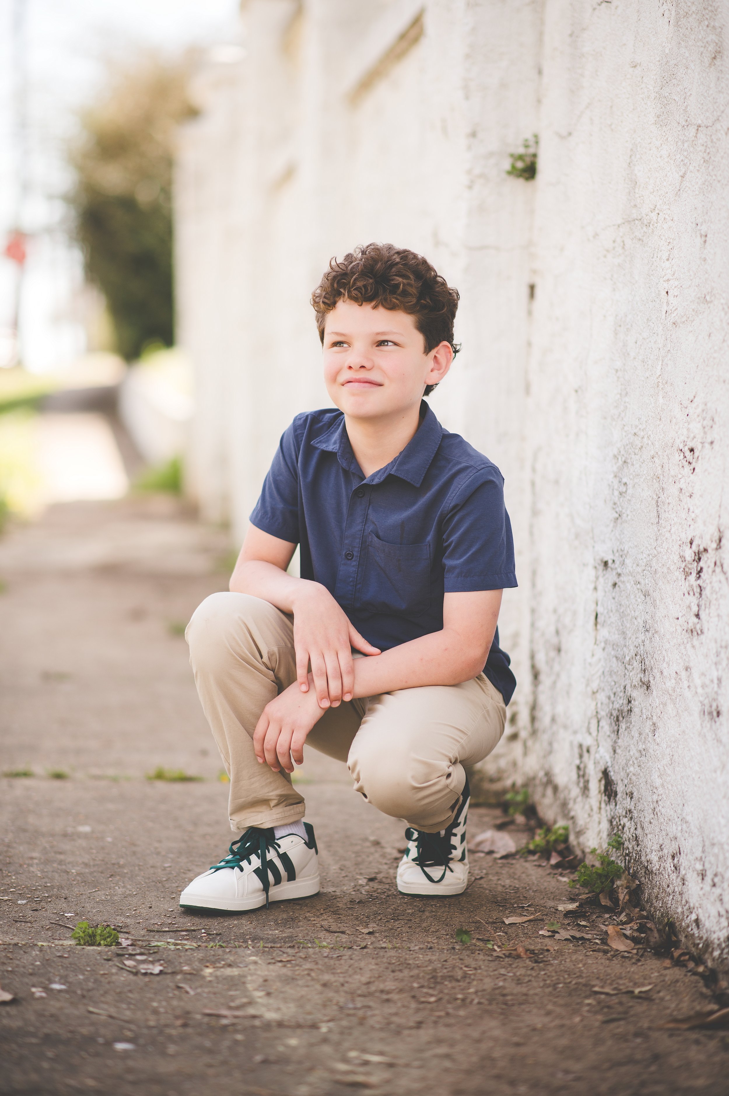 A young boy with curly brown hair, smiling, standing outdoors near train tracks with a rust-colored train car in the background, during sunset.