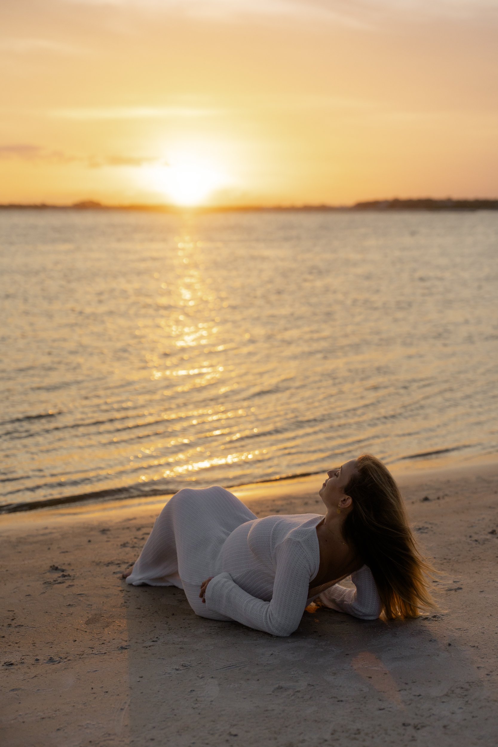 A woman in a white dress lying on the beach during sunset, with the calm water and orange sky in the background.