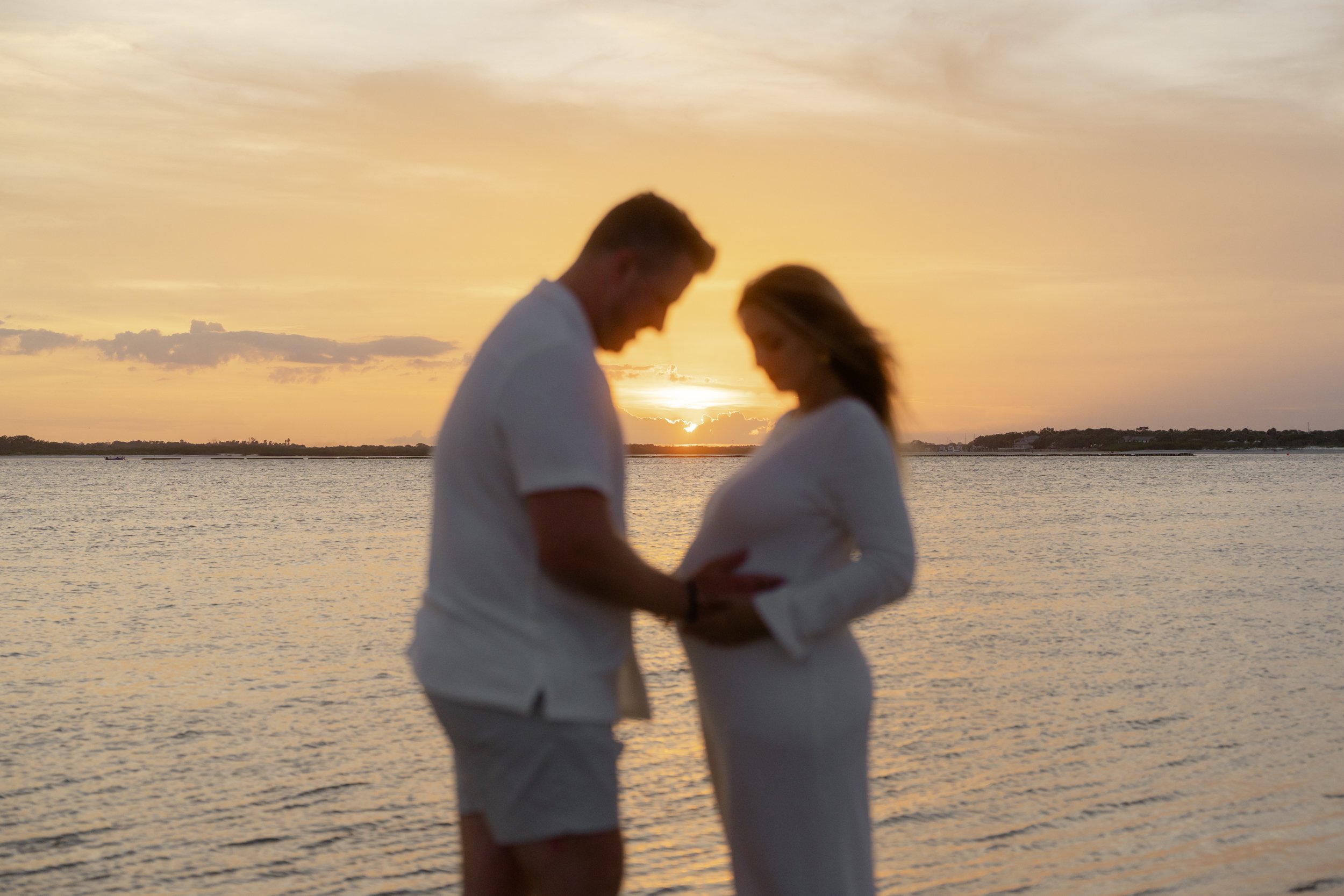 A couple, with the woman being pregnant, stands together by the water at sunset. They are looking down at the woman's belly, holding hands. The sky is filled with orange and yellow hues, reflecting on the water.