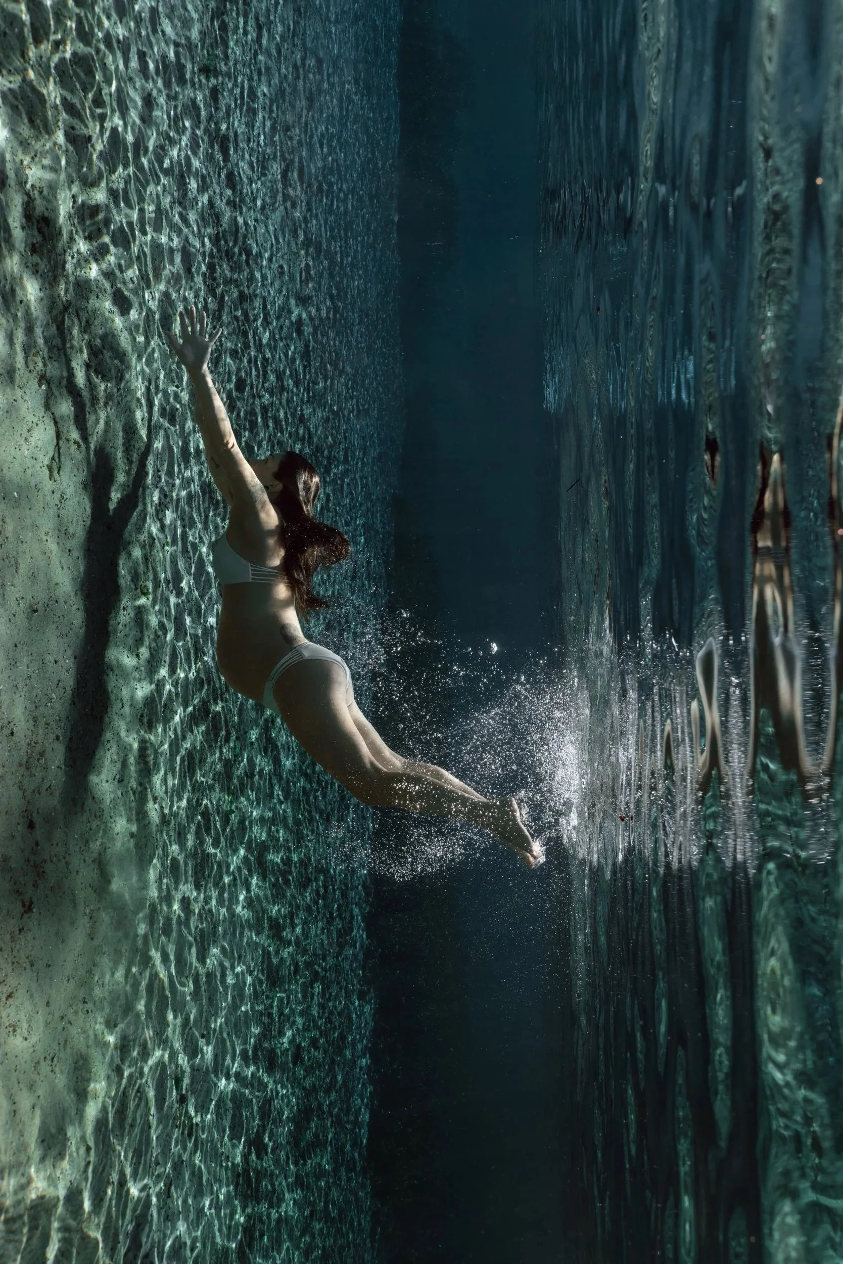 A pregnant woman in a white bikini dives into a swimming pool, creating a splash on the water's surface.