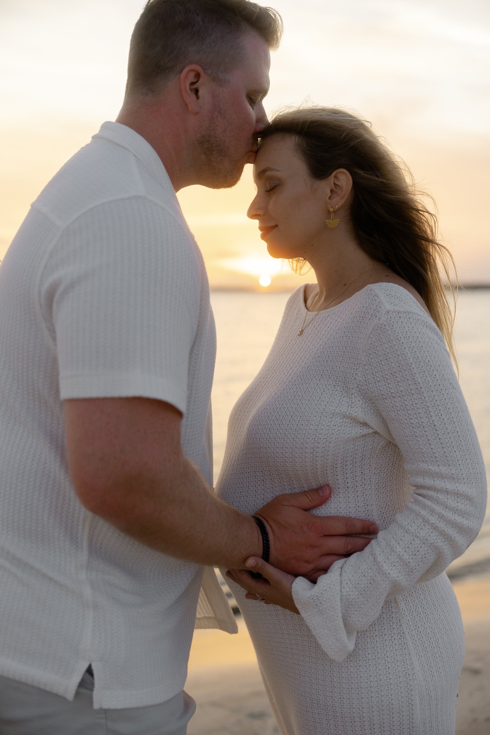 A couple stands close together on a beach at sunset, with the man kissing the woman on her forehead and the woman smiling with her eyes closed.