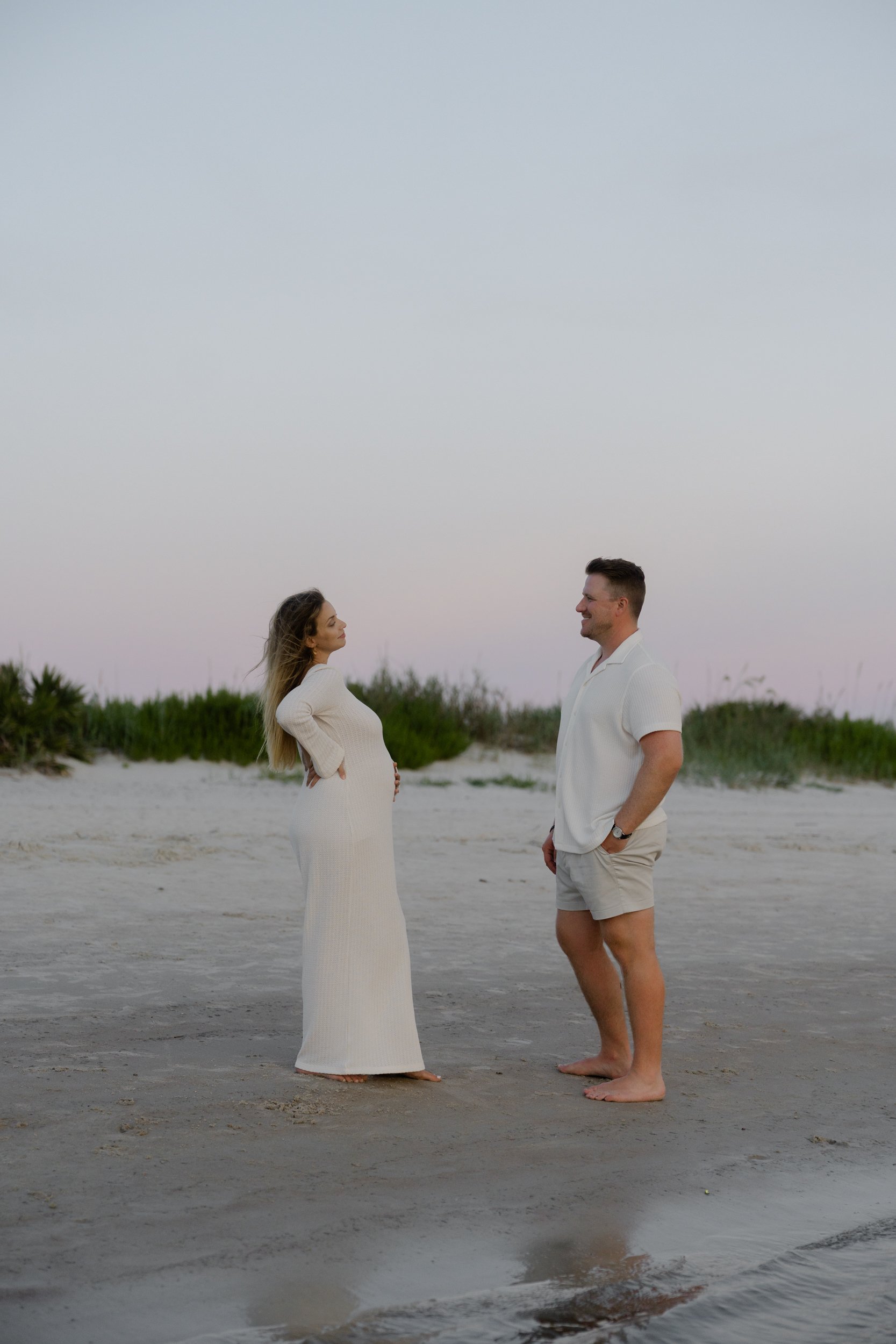 A pregnant woman and a man standing on the beach facing each other, smiling, with some greenery and a pastel sky in the background.