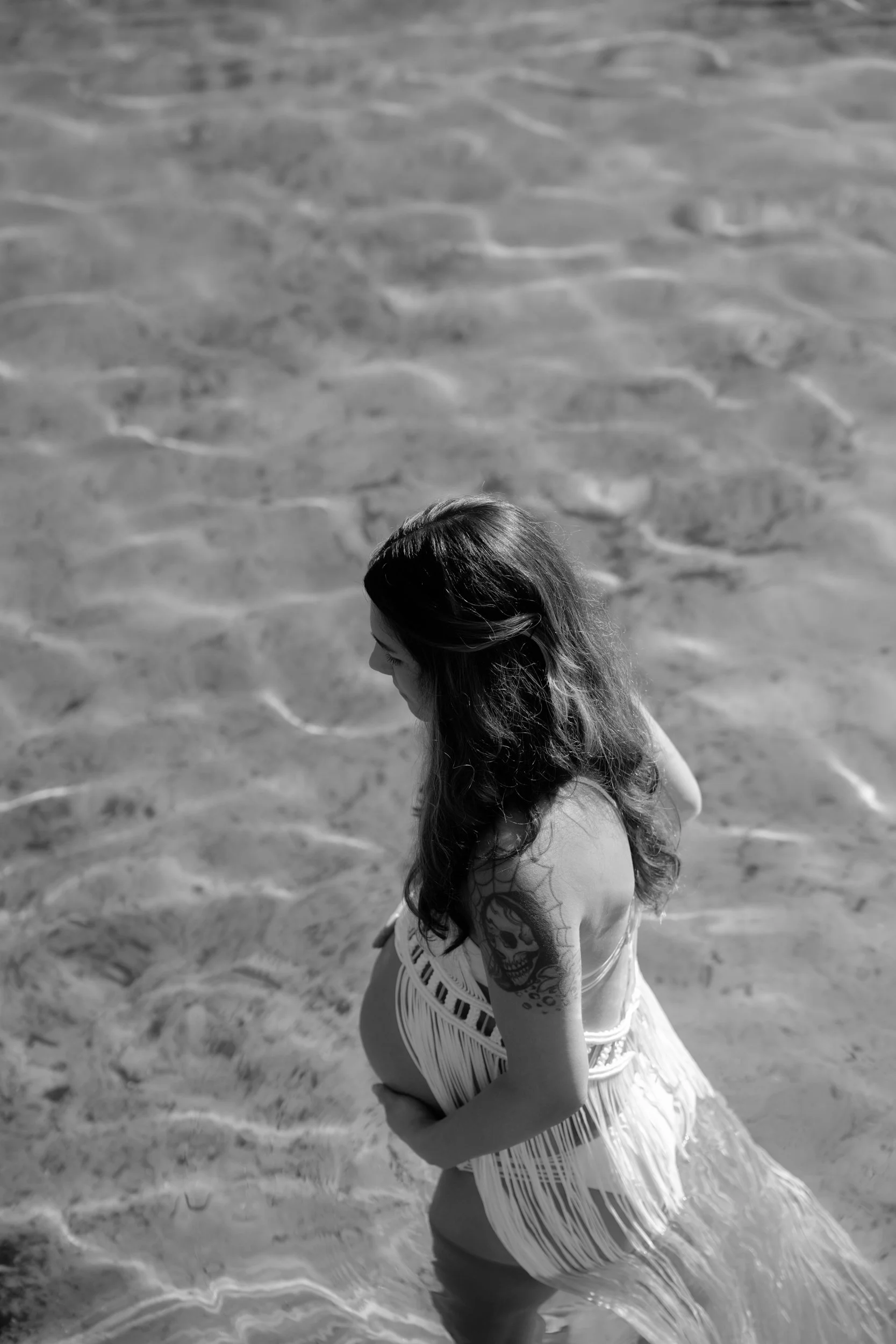 A pregnant woman stands in the shallow water of a beach, looking down with her long hair falling over her shoulder, wearing a swimsuit and a fringed cover-up, and has a tattoo of a skull on her arm.