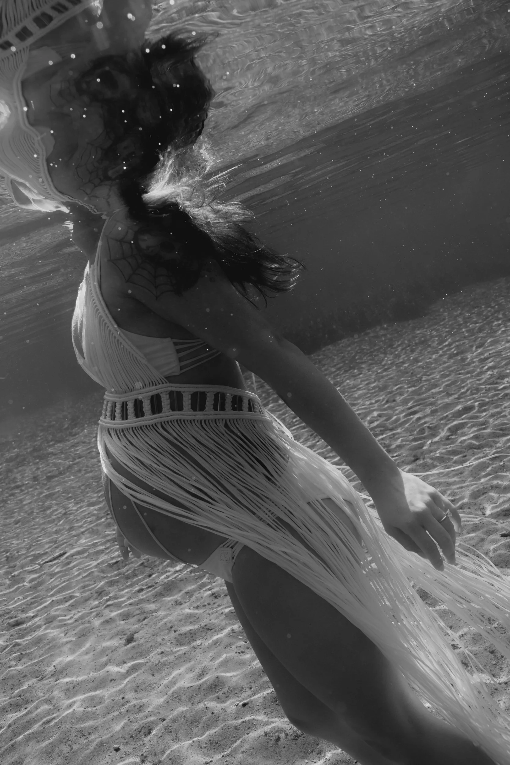 A woman underwater at the springs, wearing a swimsuit with fringes, with her hair flowing and a spiderweb tattoo on her shoulder.