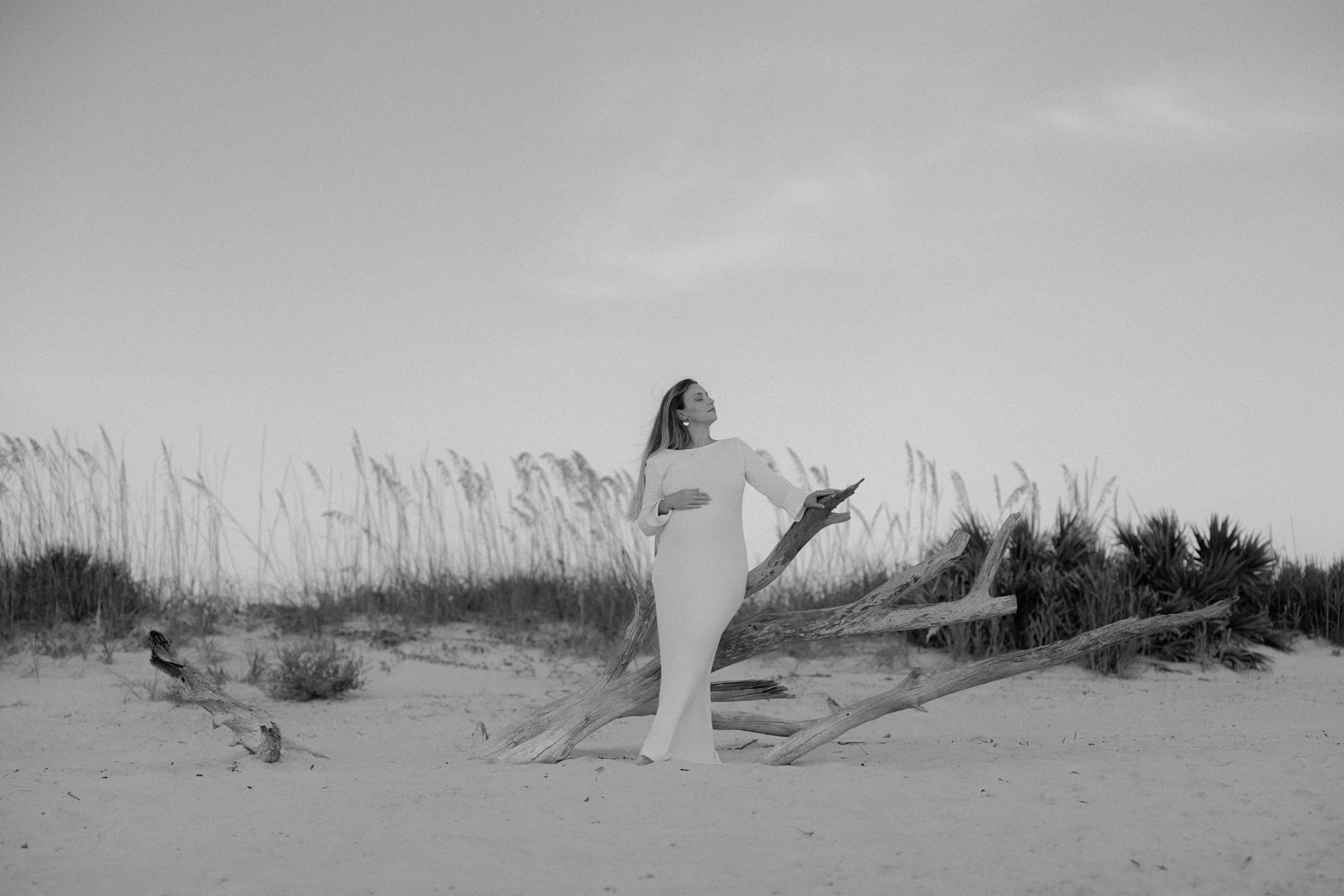 A woman in a white dress standing on a sandy beach, leaning against a driftwood branch, with tall grass and bushes in the background, captured in black and white.