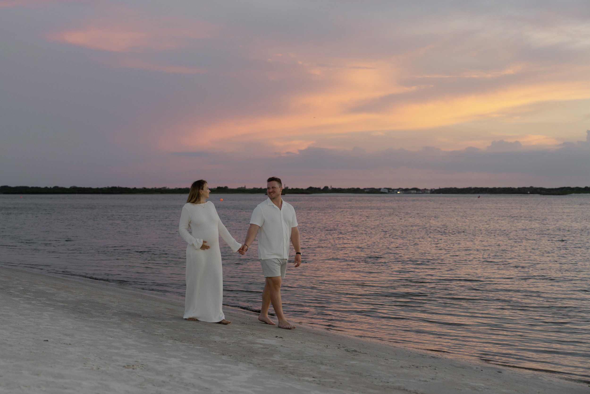 A pregnant woman and a man holding hands walking along a beach during sunset, both dressed in white, with the woman wearing a long dress and the man in shorts and a short sleeve shirt.
