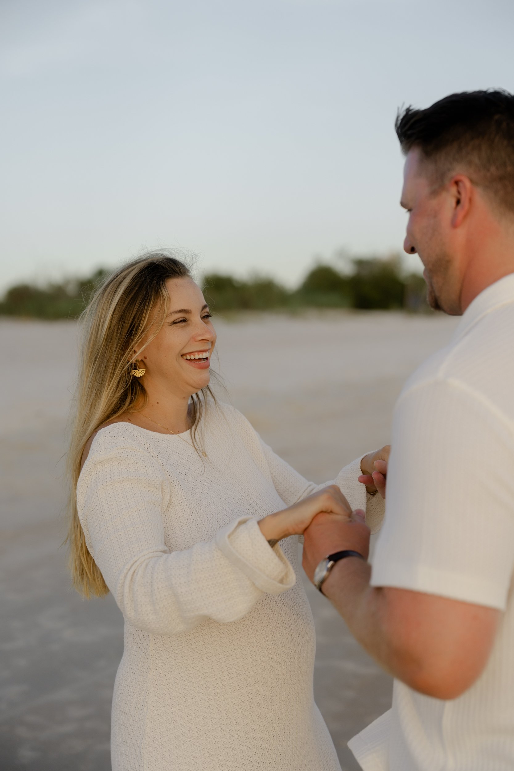 A woman with blonde hair and a white dress laughs happily while holding hands with a man on a beach during sunset.